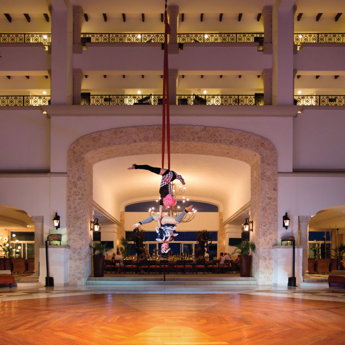 Two aerial acrobats perform on vibrant red silks in a hotel lobby, captivating wedding guests as they soar gracefully with elegant balconies in the background—a mesmerizing highlight for this unforgettable destination wedding.
