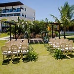 Sunny destination wedding ceremony featuring chairs arranged towards a floral arch outdoors.