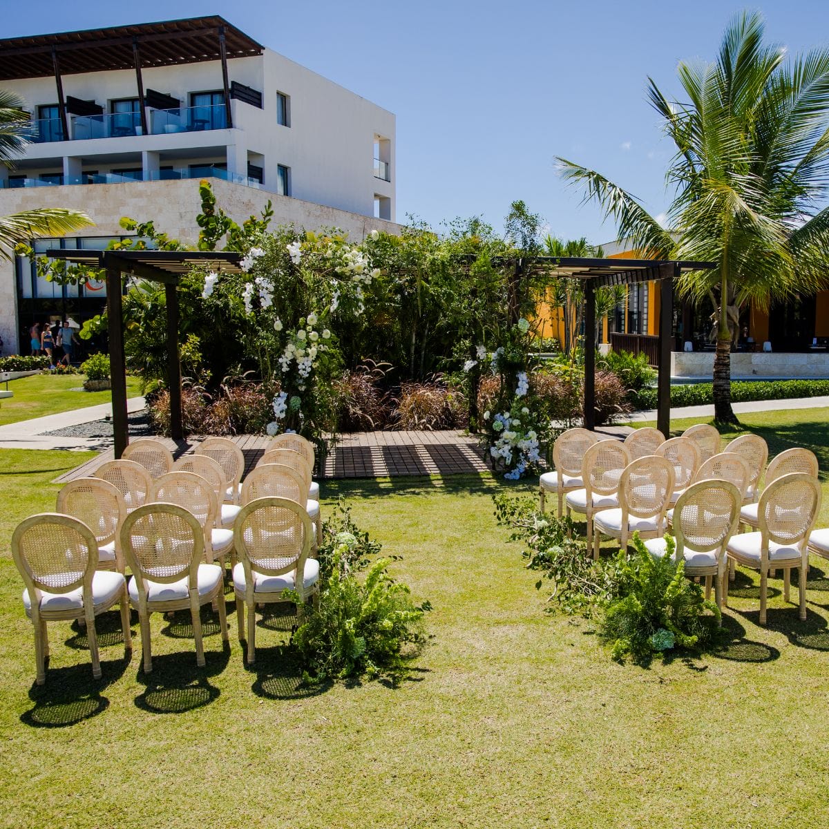 Sunny destination wedding ceremony featuring chairs arranged towards a floral arch outdoors.