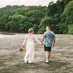 A couple holding hands walks on a sandy beach at their destination wedding, with greenery behind them.
