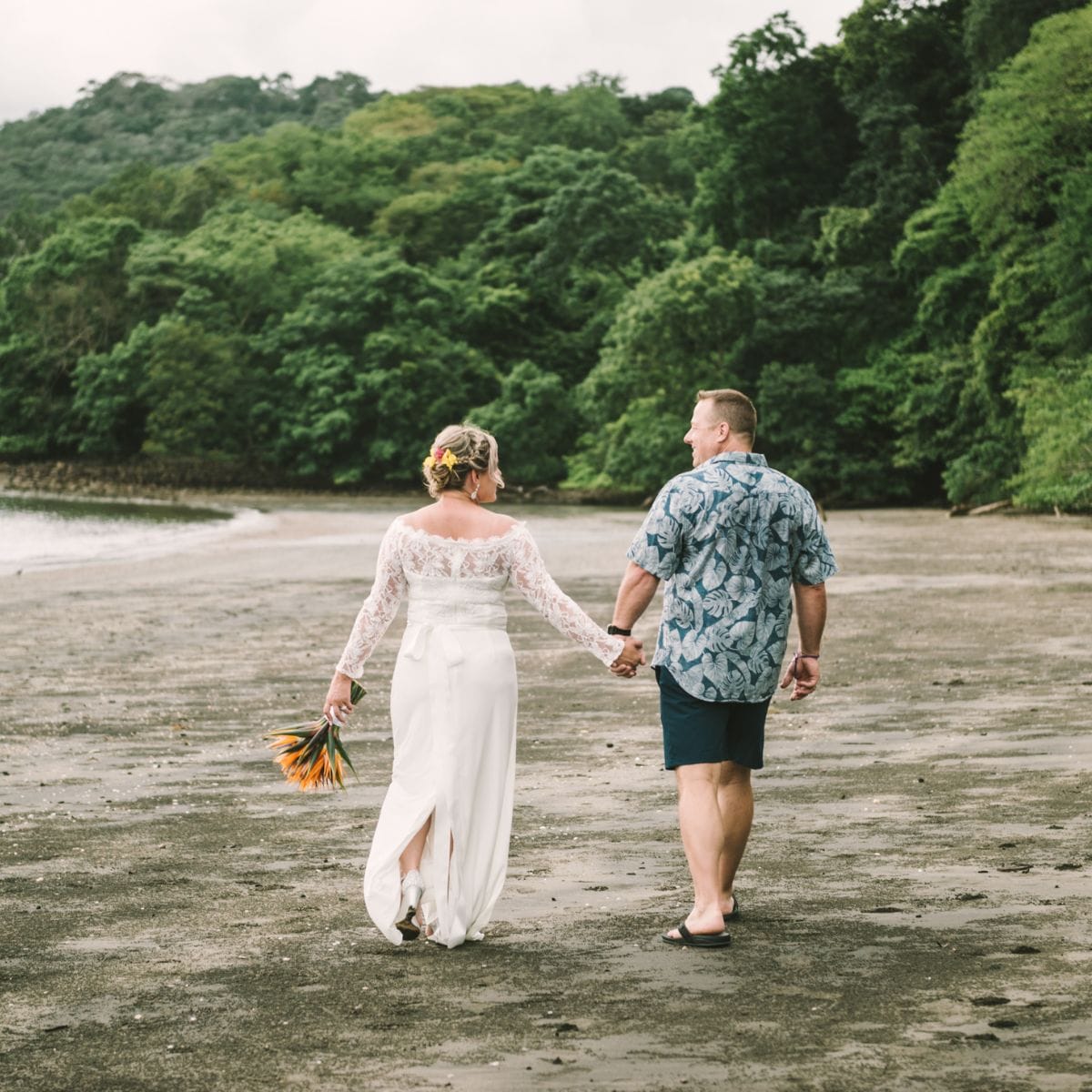 A couple holding hands walks on a sandy beach at their destination wedding, with greenery behind them.