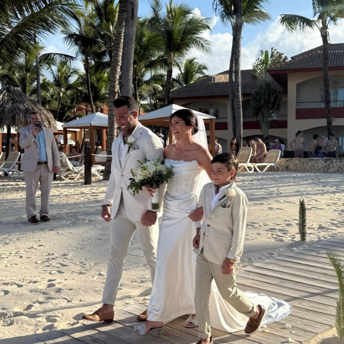 At a destination wedding, bride, groom, and young boy walk along a sandy beach during the ceremony.