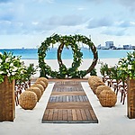 A beachfront wedding setup with wooden chairs, wicker poufs, and large green floral wreaths as the backdrop. The aisle is flanked by flower arrangements on wooden stands.