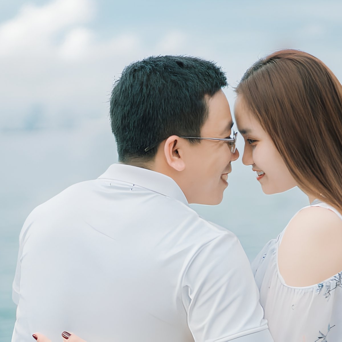 A couple sits close together by the water, smiling and touching foreheads as they celebrate their destination wedding.
