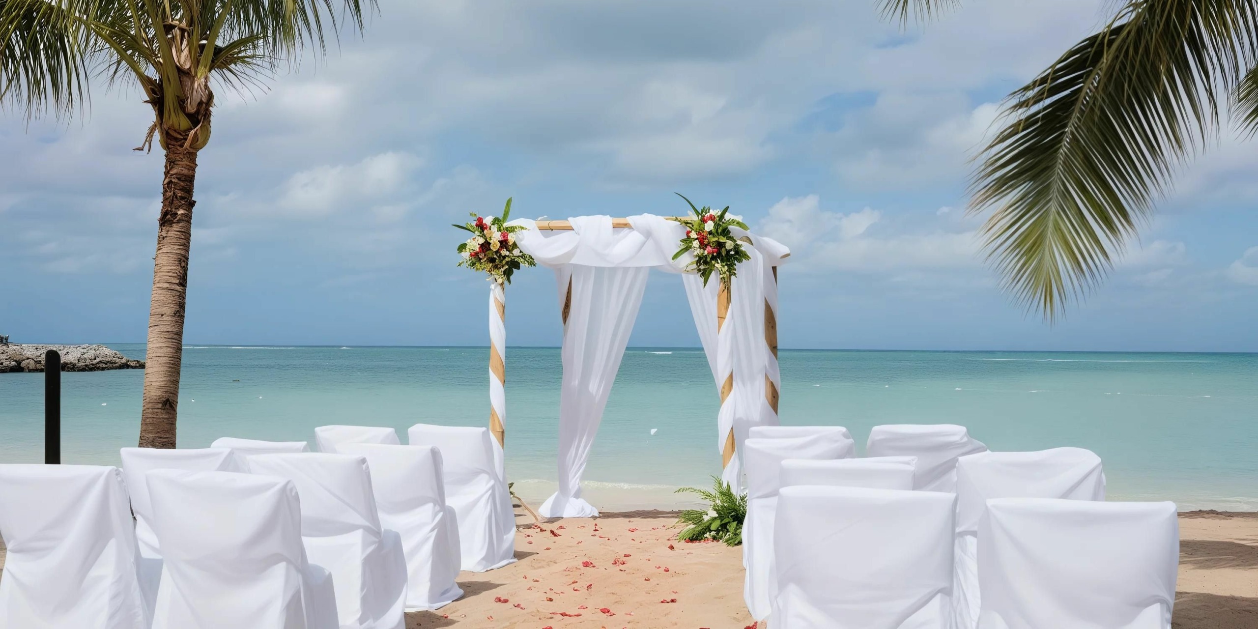 A beach wedding setup featuring a decorated arch with white fabric and flowers, rows of white-covered chairs, all in front of a calm ocean backdrop with palm trees.