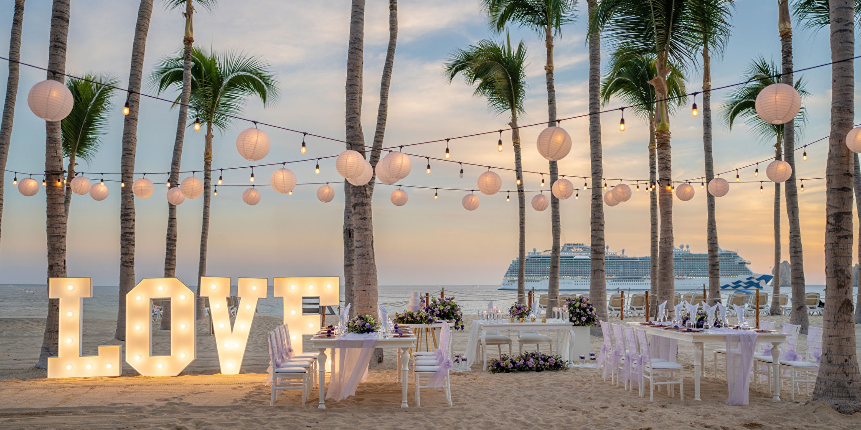 Destination wedding setup on the beach with "LOVE" marquee lights, string lanterns, and elegant tables at sunset.