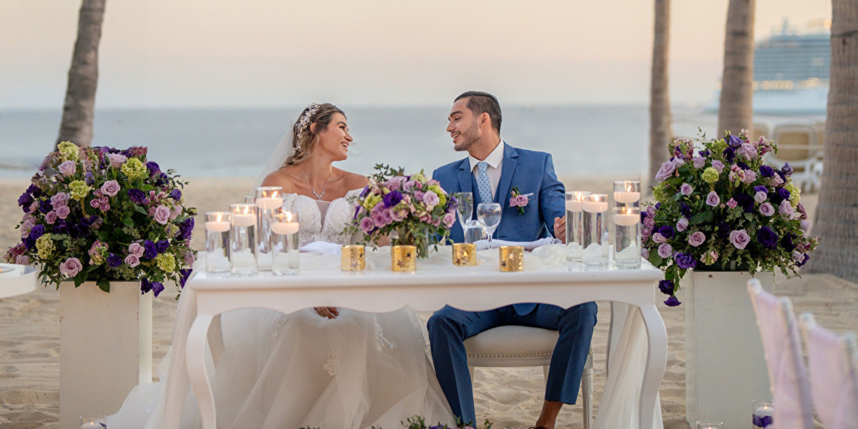 At a destination wedding, the bride and groom sit at a candlelit, purple-flowered table on the beach.