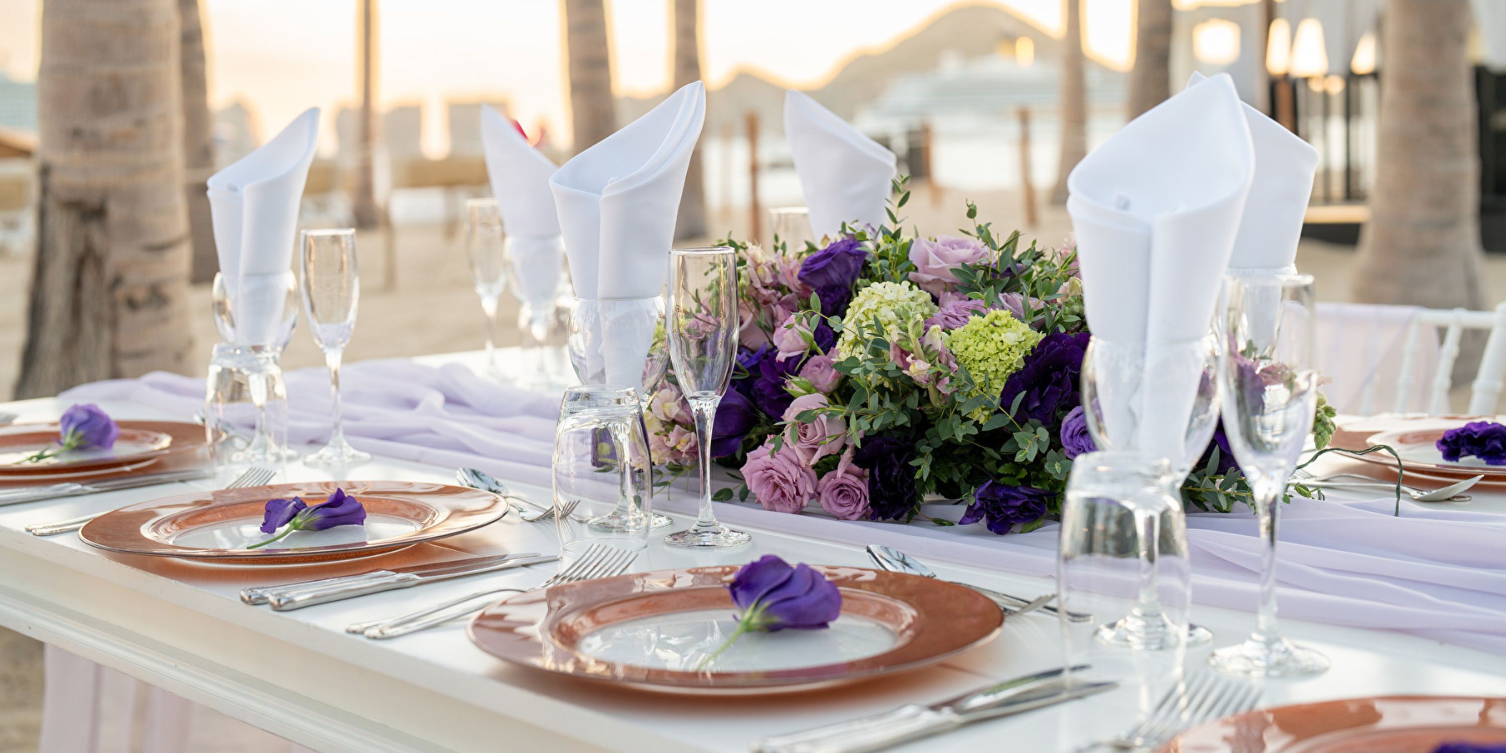 Outdoor table elegantly arranged with plates, glasses, and a purple floral centerpiece for a wedding.