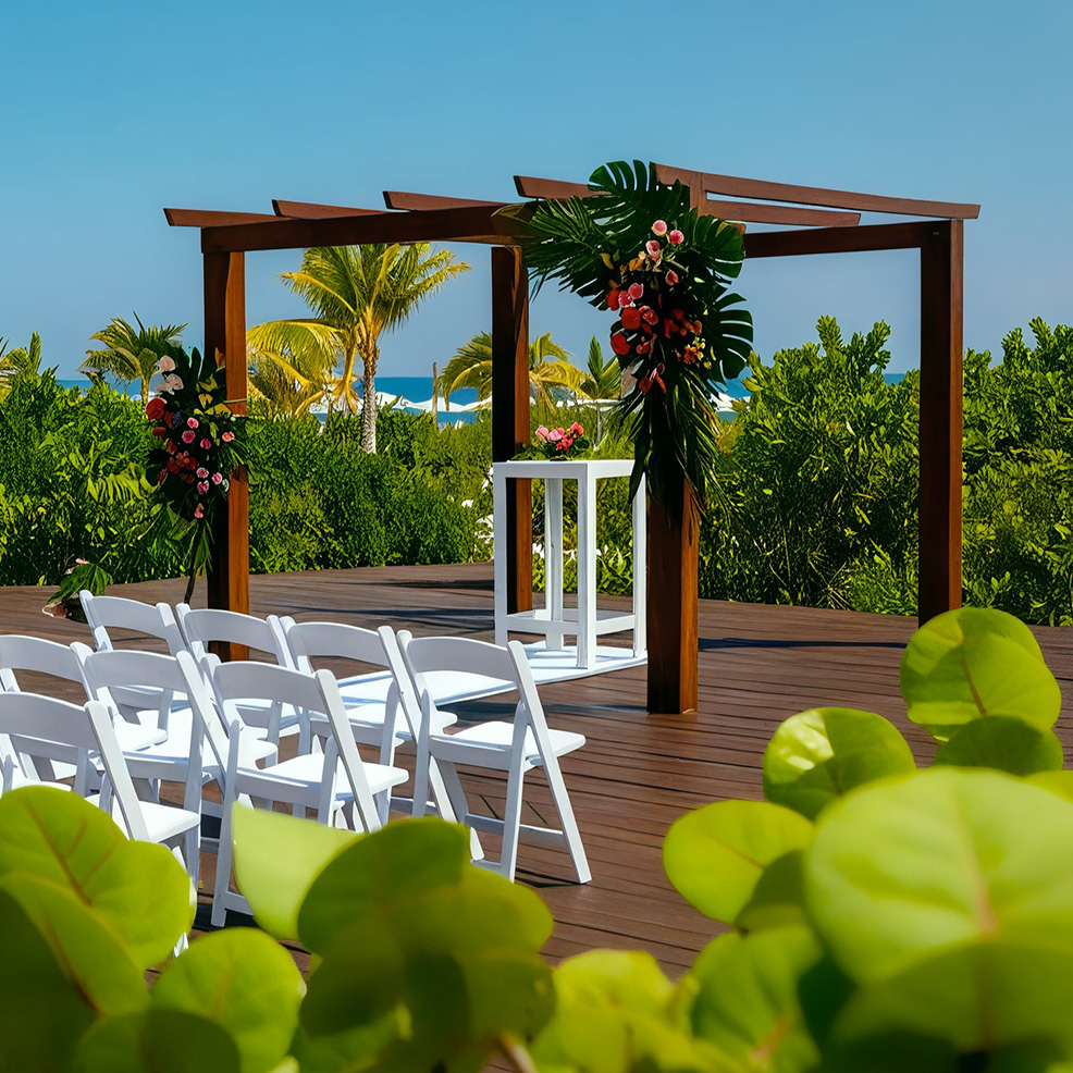 Outdoor wedding setup with white chairs facing a wooden pergola decorated with tropical flowers. Lush greenery and a glimpse of the ocean in the background.