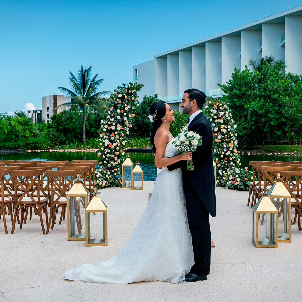 A bride and groom stand together holding flowers in an outdoor wedding setting with decorative floral arches and lanterns.