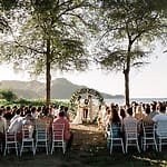 Destination wedding ceremony at a Costa Rica resort, couple stands beneath floral arch as guests look on.