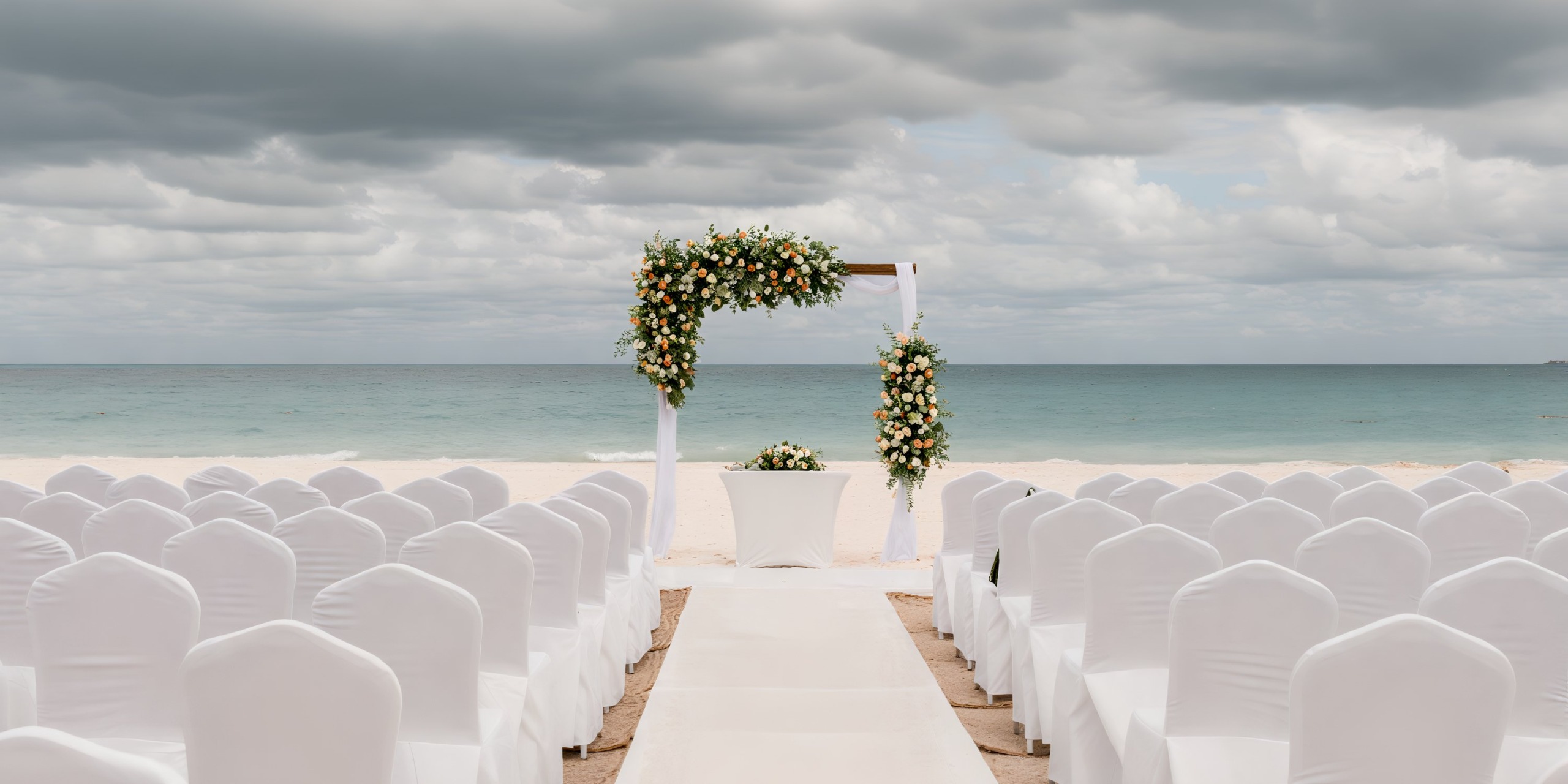 Under the cloudy skies, a majestic elegance unfolds, showcasing a beautifully arranged wedding setup with chairs and a floral arch.