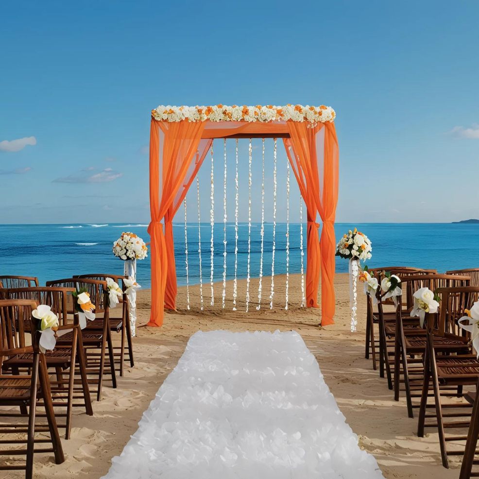 A beach wedding setup with wooden chairs, white flowers on stands, an orange draped arch with flower garlands, and a white aisle runner leading to the arch by the ocean.