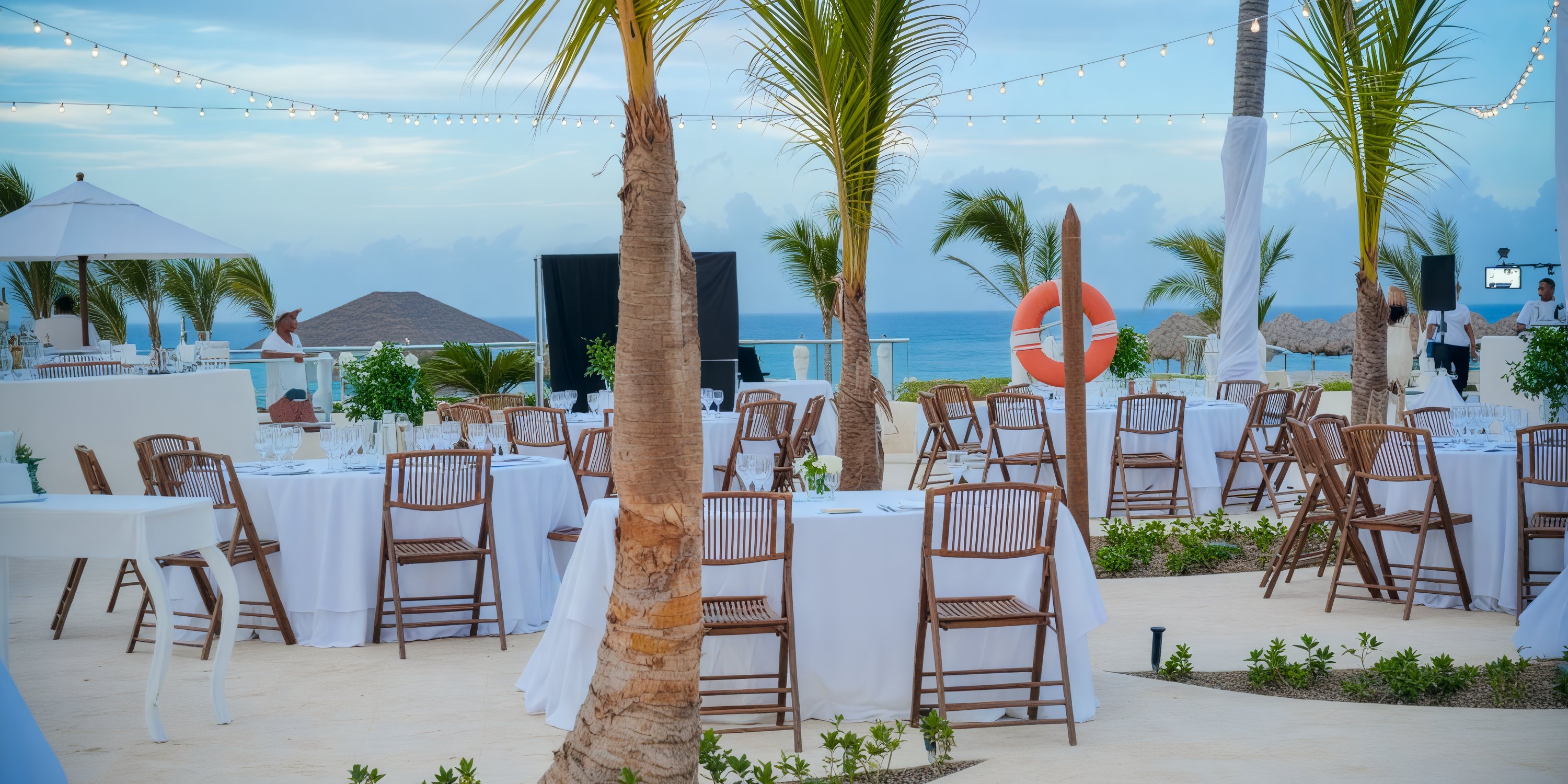 Experience a destination wedding at Breathless Punta Cana featuring a beachside event elegantly arranged with round tables, adorned with white cloths, and complemented by charming wicker chairs.