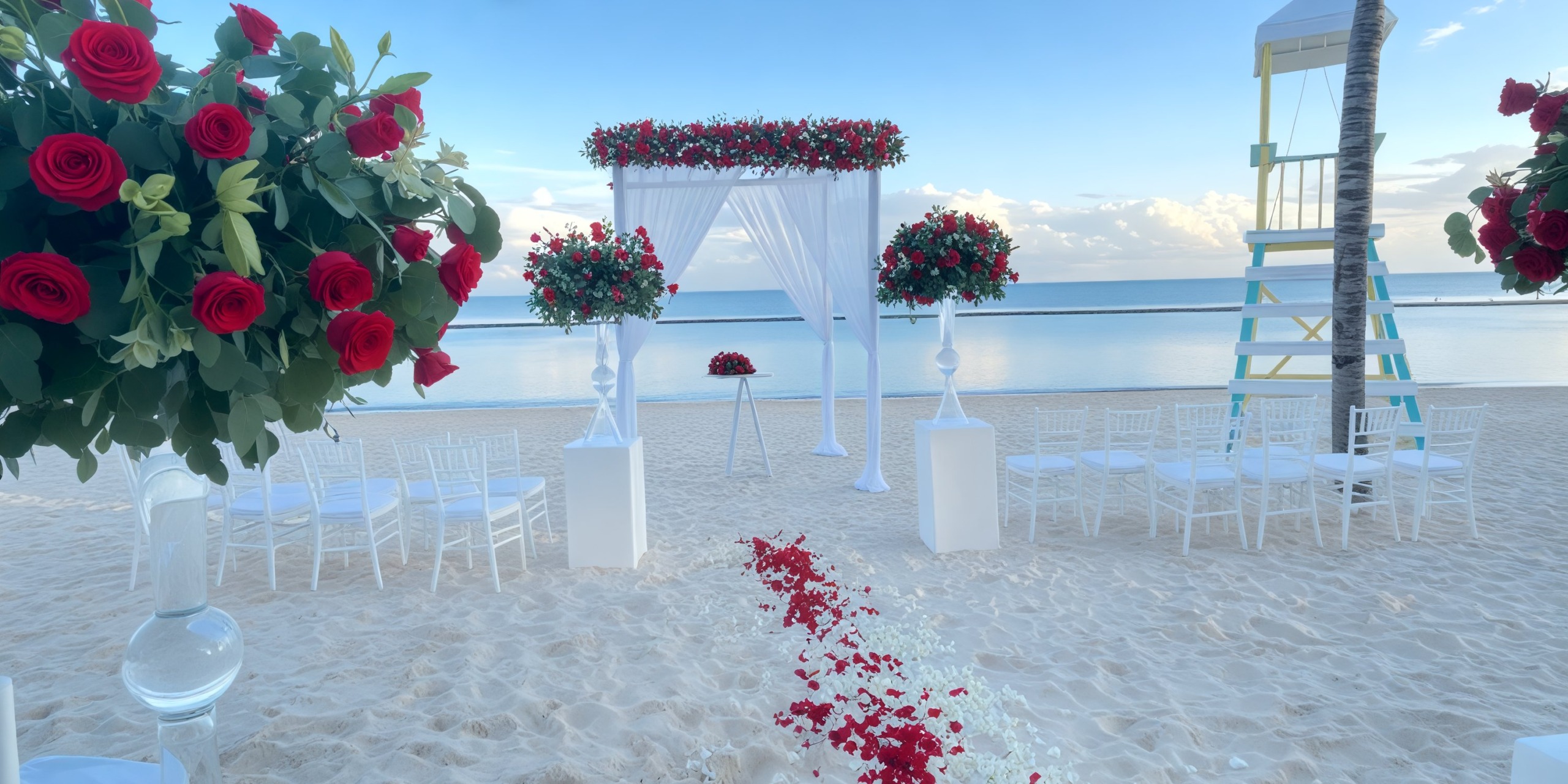 A picturesque beachfront destination wedding setup featuring red roses and elegant white chairs all arranged to face a stunning floral arch.
