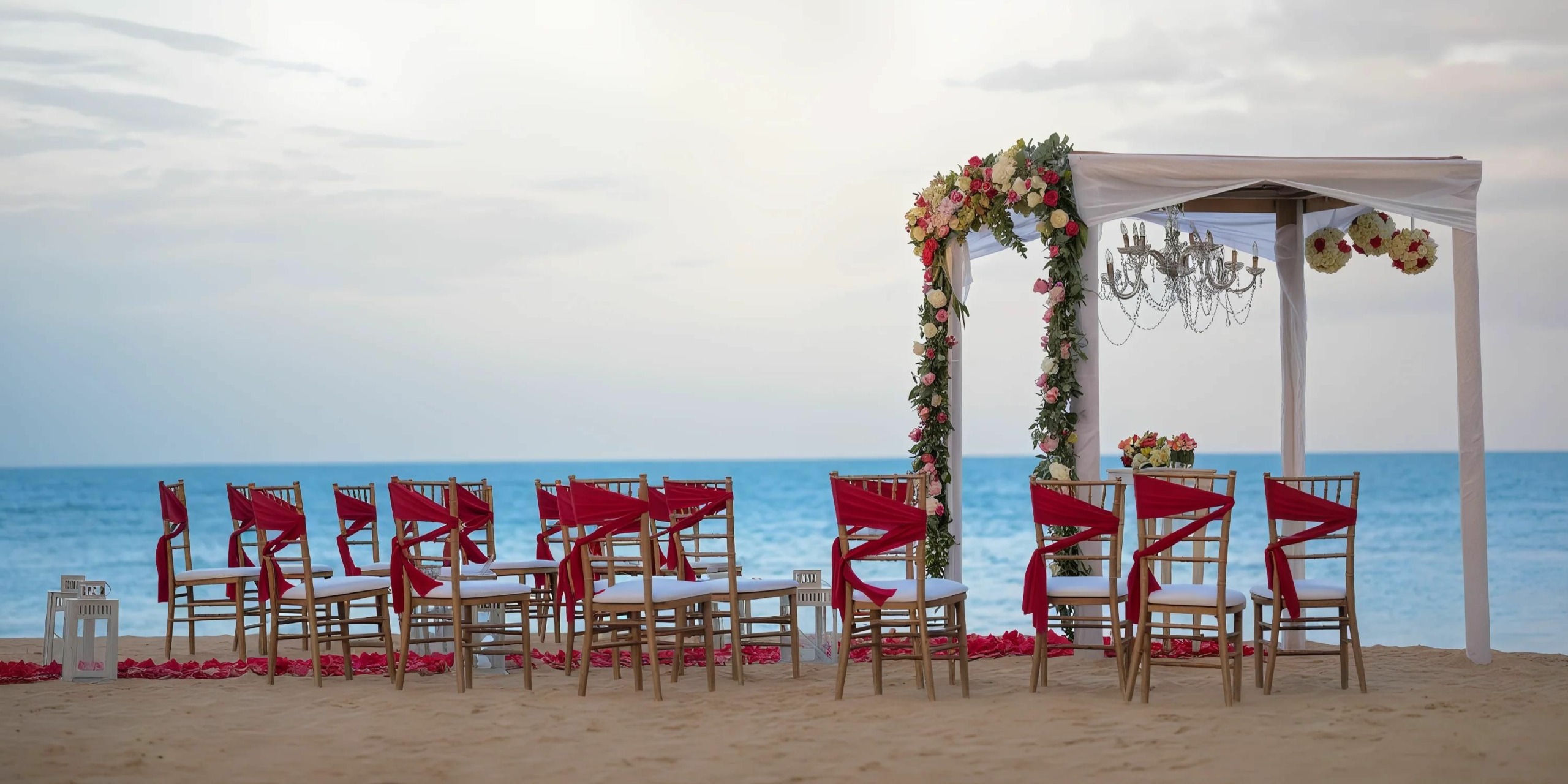 A beach wedding setup at Breathless Punta Cana Resort and Spa features rows of wooden chairs adorned with red sashes, all facing the serene ocean. At the front stands a beautifully decorated canopy, elegantly draped with flowers and complemented by a chandelier.