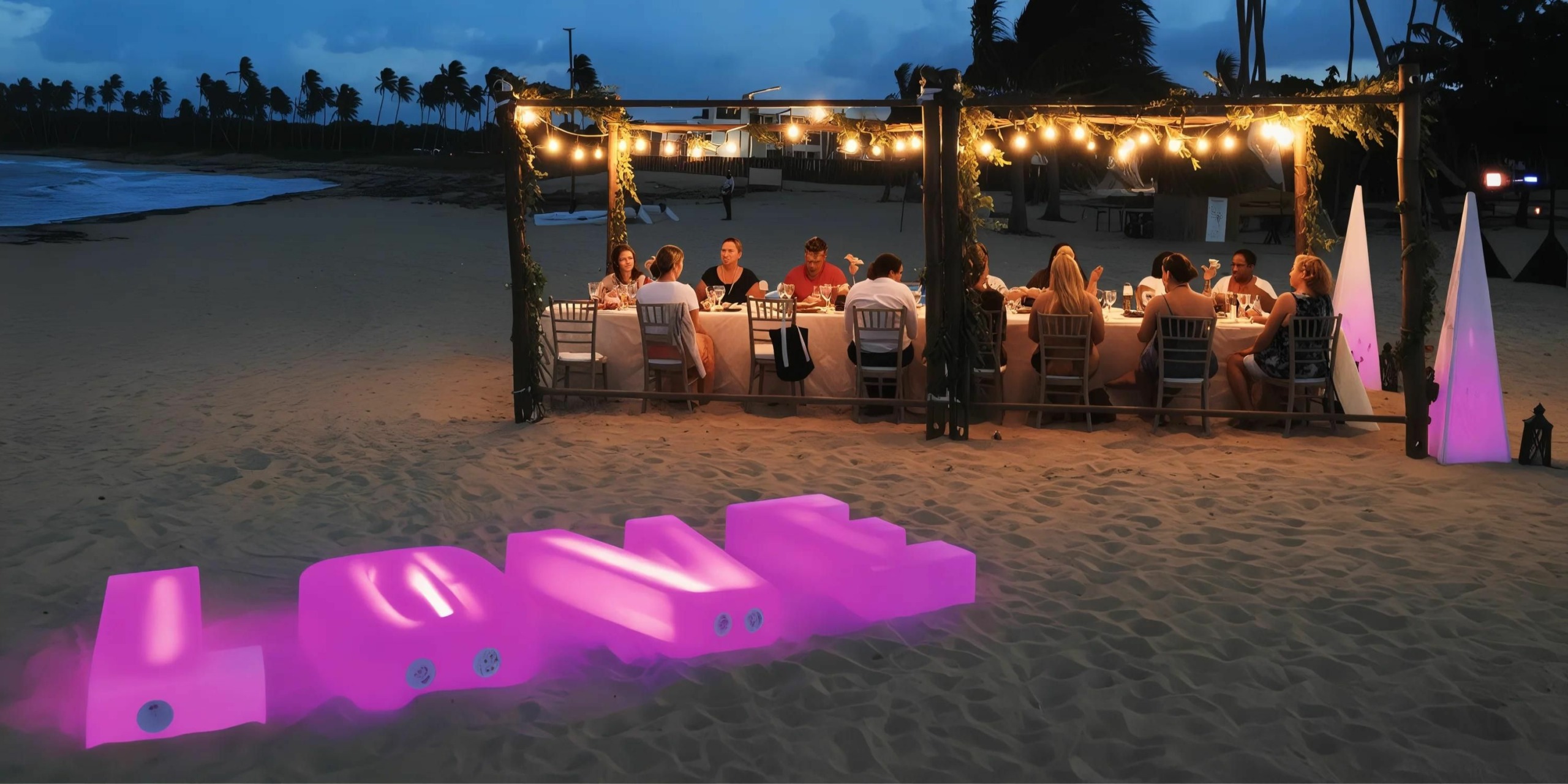 A group of people dining at a long table on the sandy beach of Breathless Punta Cana Resort and Spa, illuminated by string lights at dusk, with glowing 'LOVE' letters displayed in the foreground.