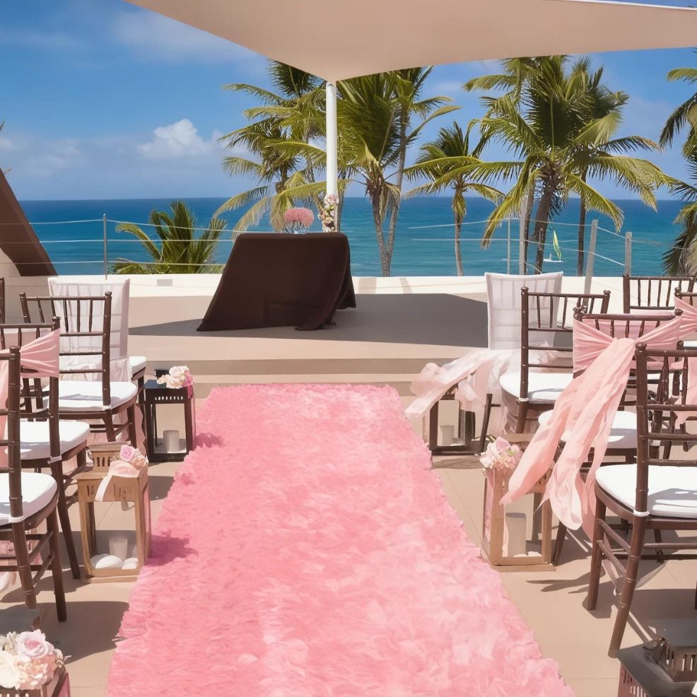 An outdoor wedding ceremony setup with a pink aisle runner, chairs with pink sashes, and a small stage under a white canopy overlooking the ocean and palm trees.