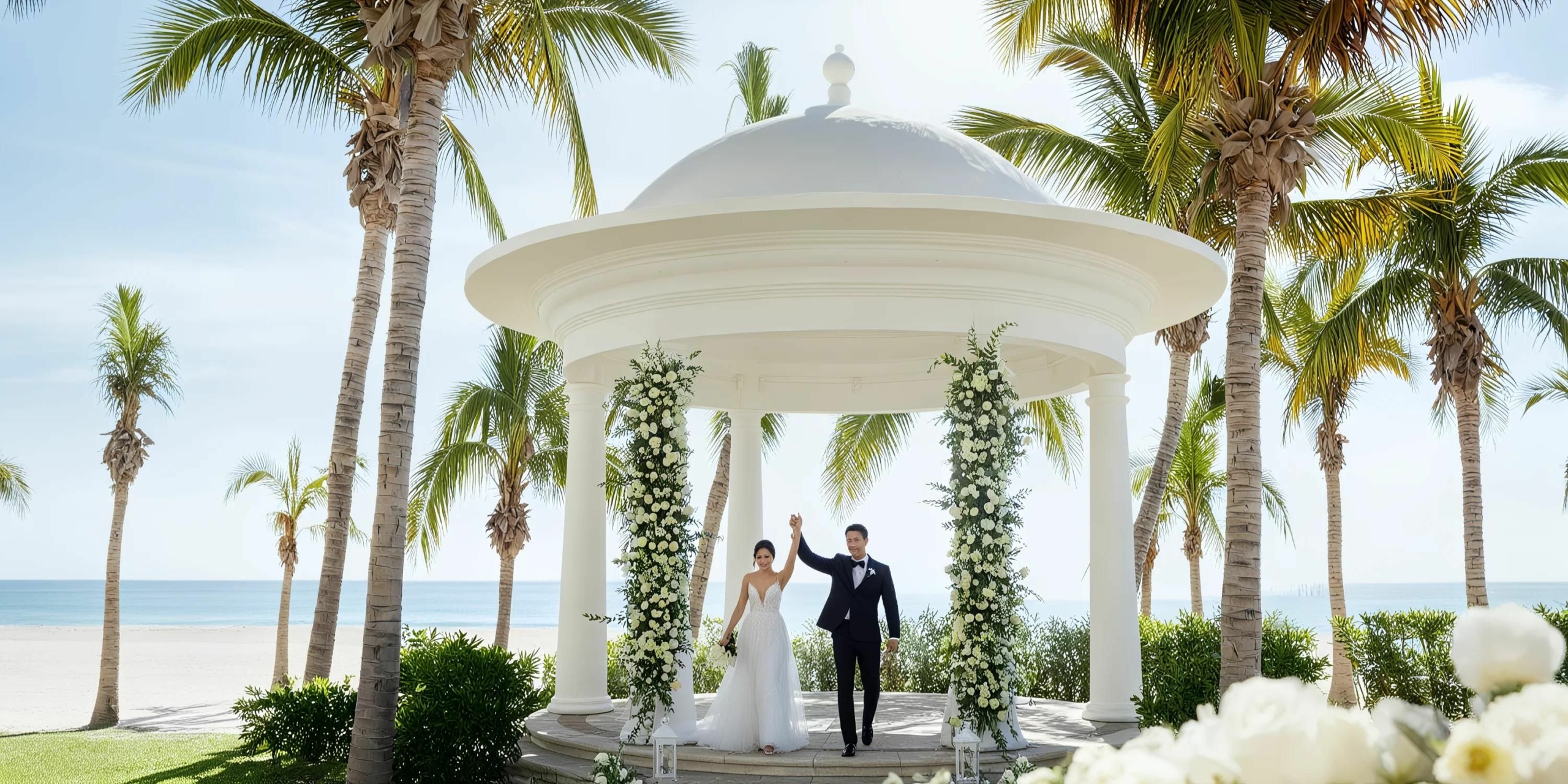A bride and groom celebrate under a white gazebo at the Hyatt Ziva Los Cabos, surrounded by palm trees on a beach, on a sunny day.