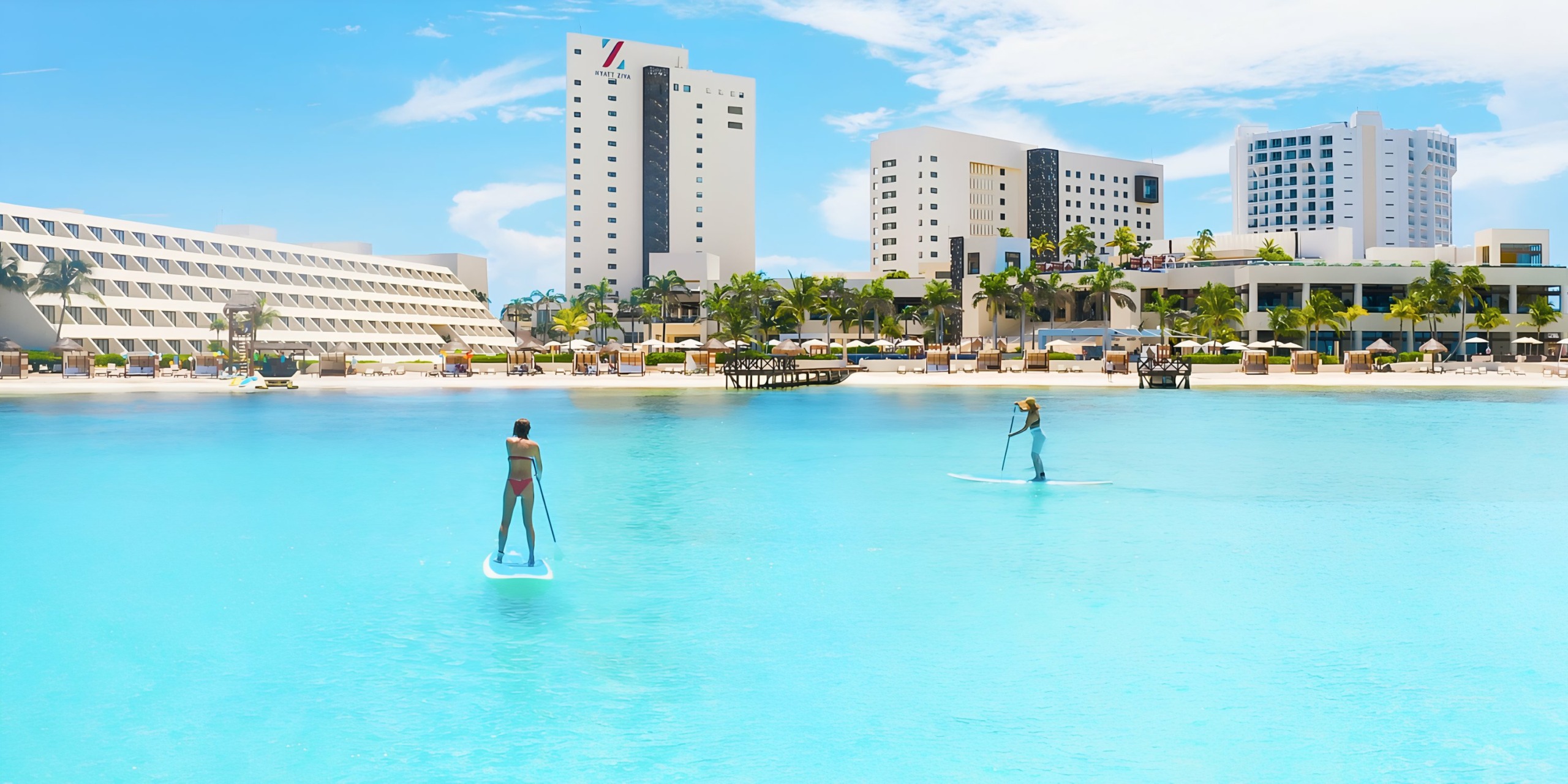 Two people paddleboarding on a calm turquoise sea near Hyatt Ziva Cancun with white buildings.