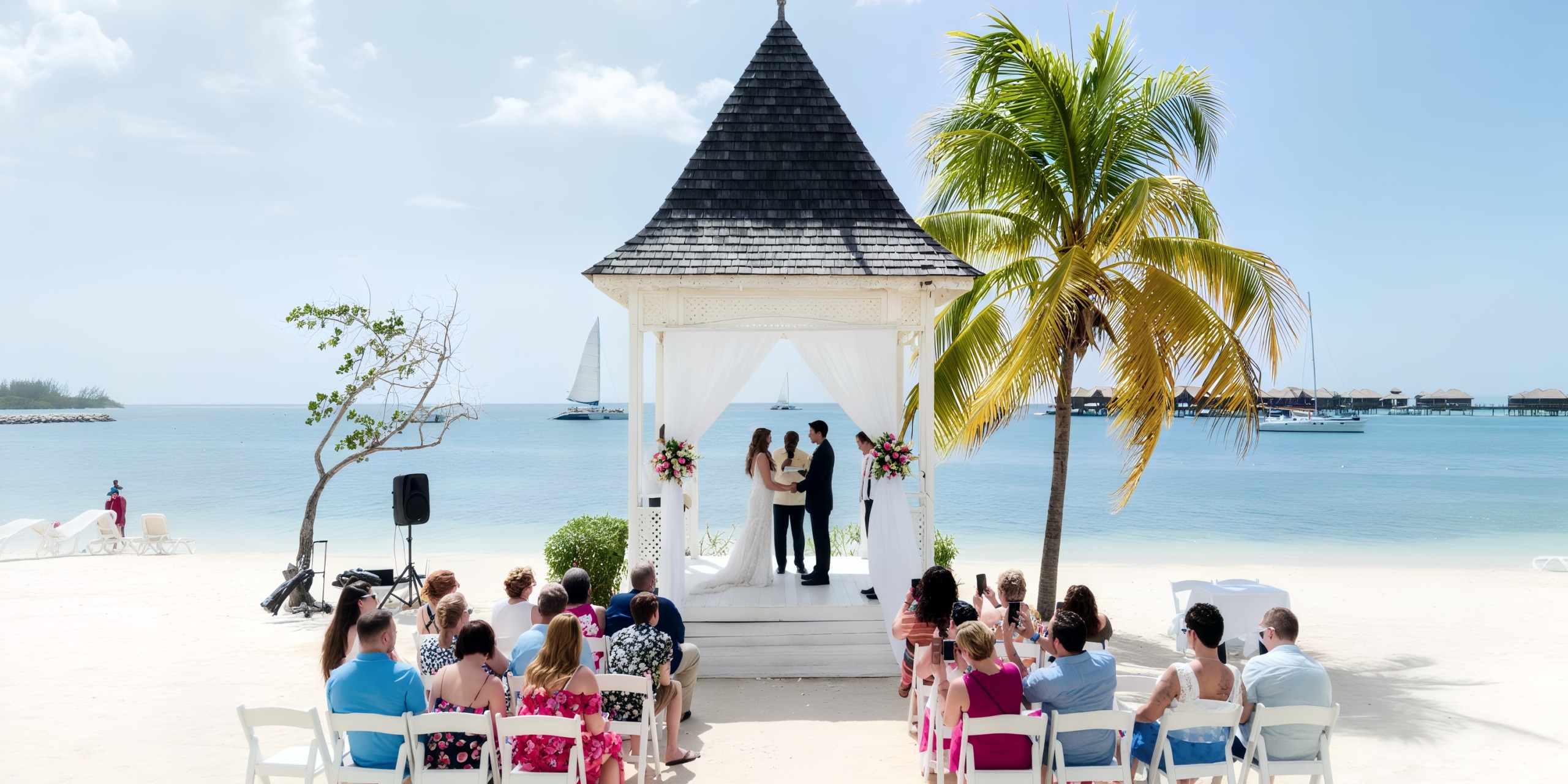 A couple exchanges vows under a gazebo on the beach at Hotel Riu Montego Bay, surrounded by guests seated in chairs. Nearby, a palm tree adds to the tropical ambiance, and a sailboat can be seen gliding across the water.