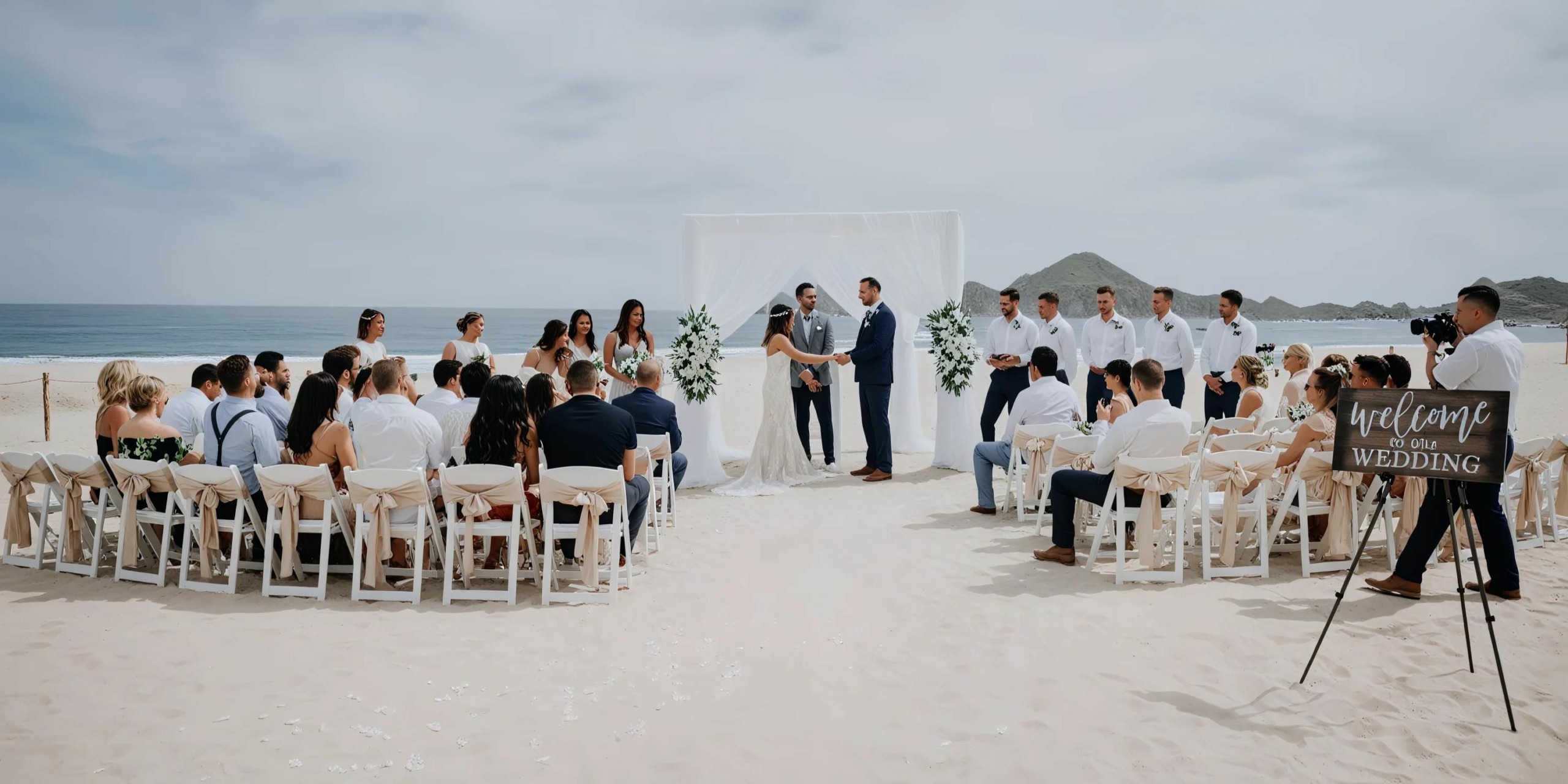 A beach wedding ceremony at Hotel Riu Palace Cabo San Lucas, with guests seated in rows facing the ocean. The couple stands under an arch with the officiant. A sign reads "Welcome to Our Wedding." Groomsmen and bridesmaids are on either side.