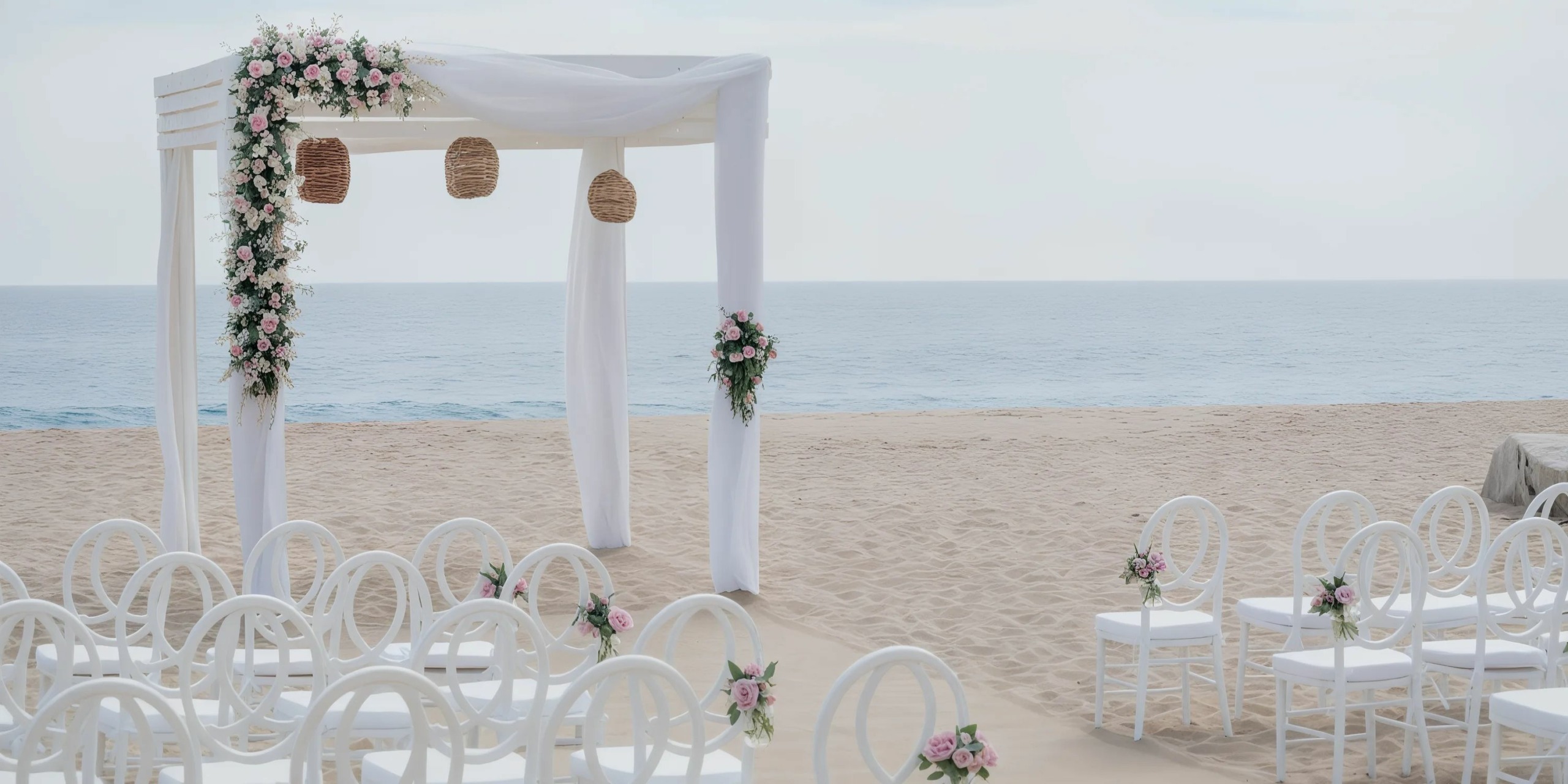 Romantic beach wedding setup at Sandos Finisterra Los Cabos featuring a white pergola embellished with pink and white flowers, hanging wicker lanterns, and rows of white chairs overlooking the ocean.