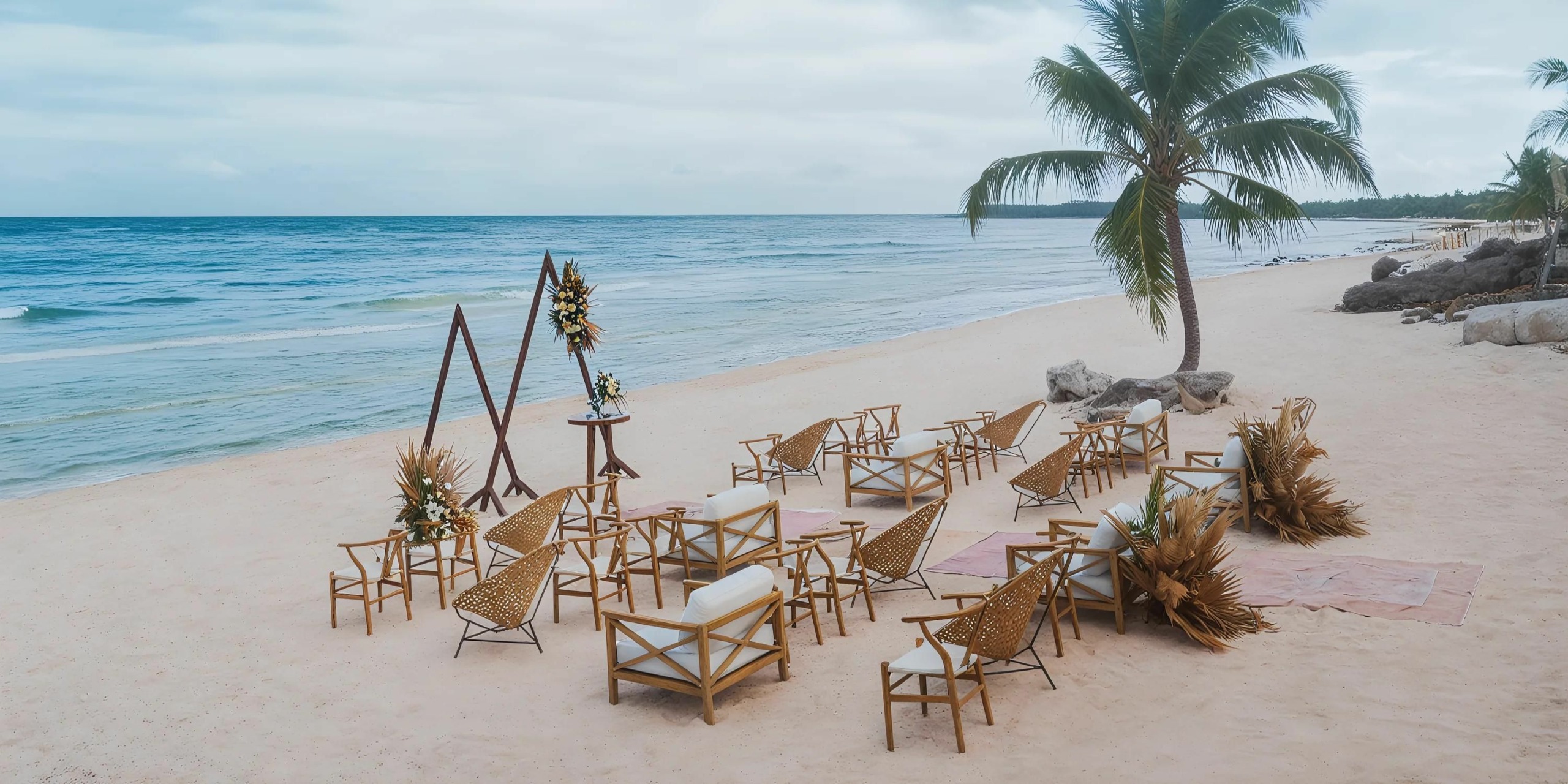 A beachside wedding setup at UNICO 20°87° features chairs elegantly arranged in a semi-circle facing a beautifully decorated archway adorned with vibrant flowers. A majestic palm tree stands in the background, and the gentle ocean waves are visible, creating a picturesque scene.