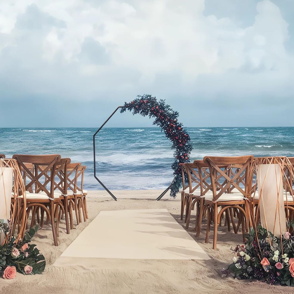 A beach wedding setup with wooden chairs arranged in two rows facing a simple geometric arch adorned with flowers, set against a backdrop of ocean waves and a cloudy sky.