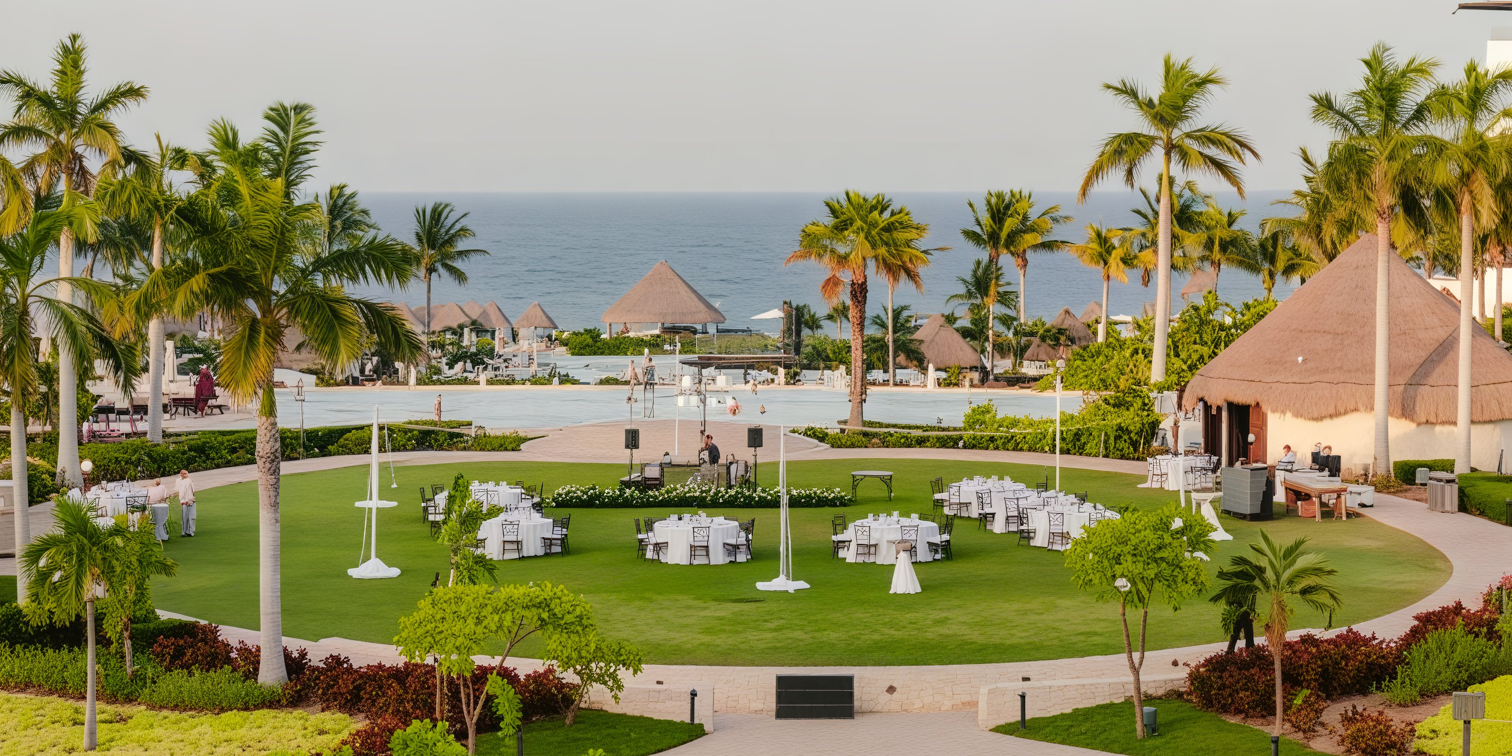 A beachfront resort perfect for a destination wedding, featuring a pool, swaying palm trees, and a charming round dining setup on a lush grassy area.