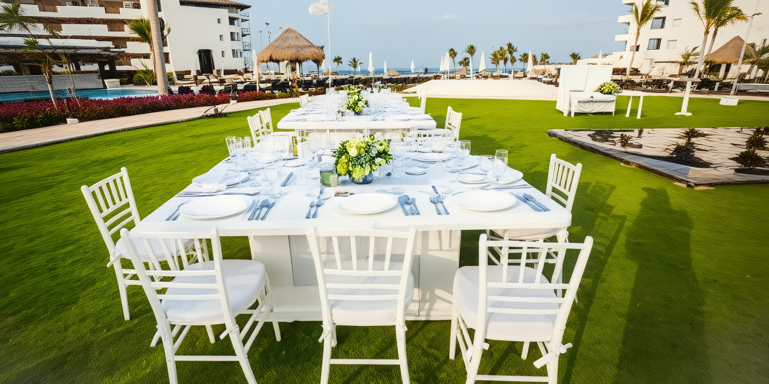 White tables and chairs, elegantly arranged with dishes and flowers, grace the green lawn beside a poolside setting, creating a stunning scene for a destination wedding.