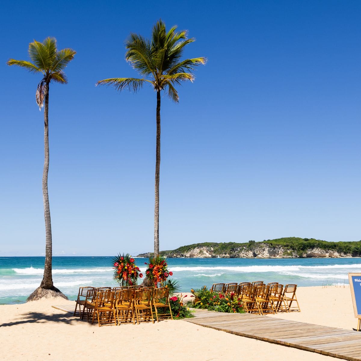 Rows of chairs and floral arrangements arranged for a destination wedding on a sandy beach with palm trees.