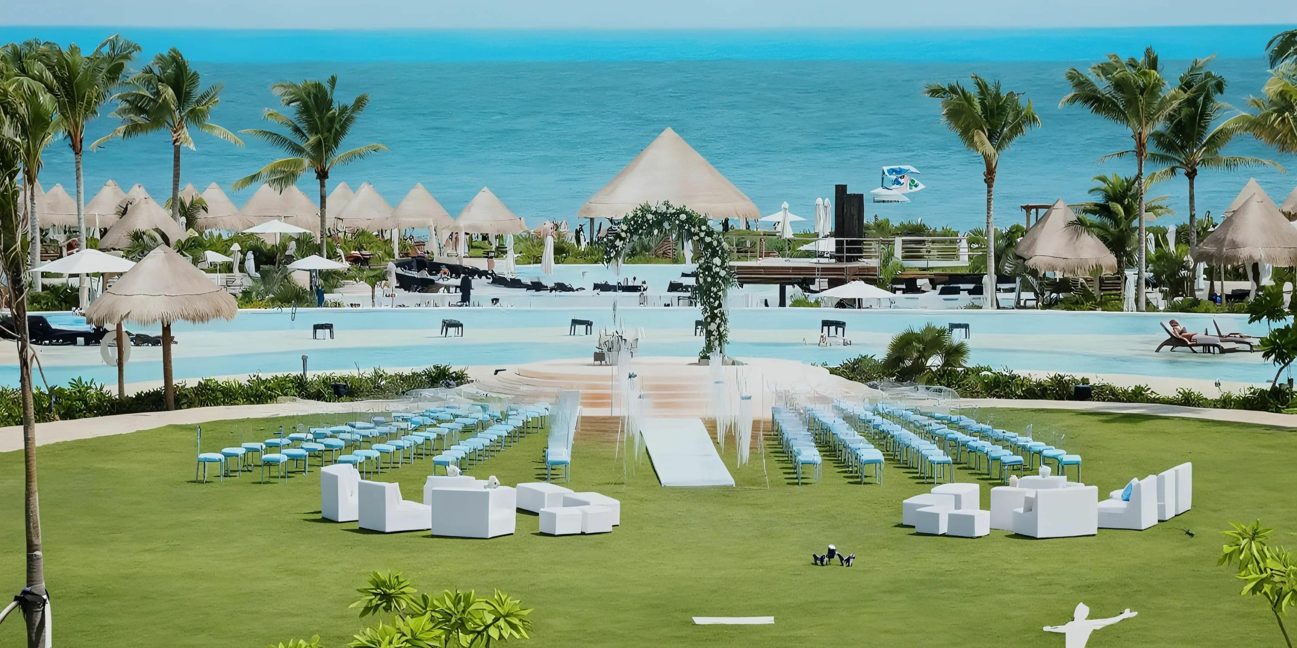 Outdoor wedding setup on a lawn with white chairs and a floral arch overlooking a pool and the ocean, surrounded by palm trees and cabana-style structures.