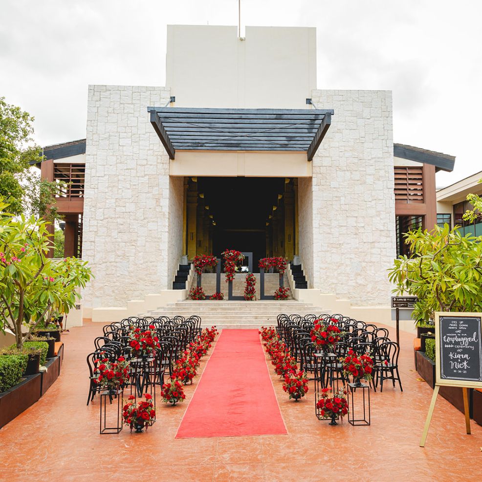 Destination wedding ceremony featuring a red carpet aisle, black chairs, and elegant red floral arrangements.