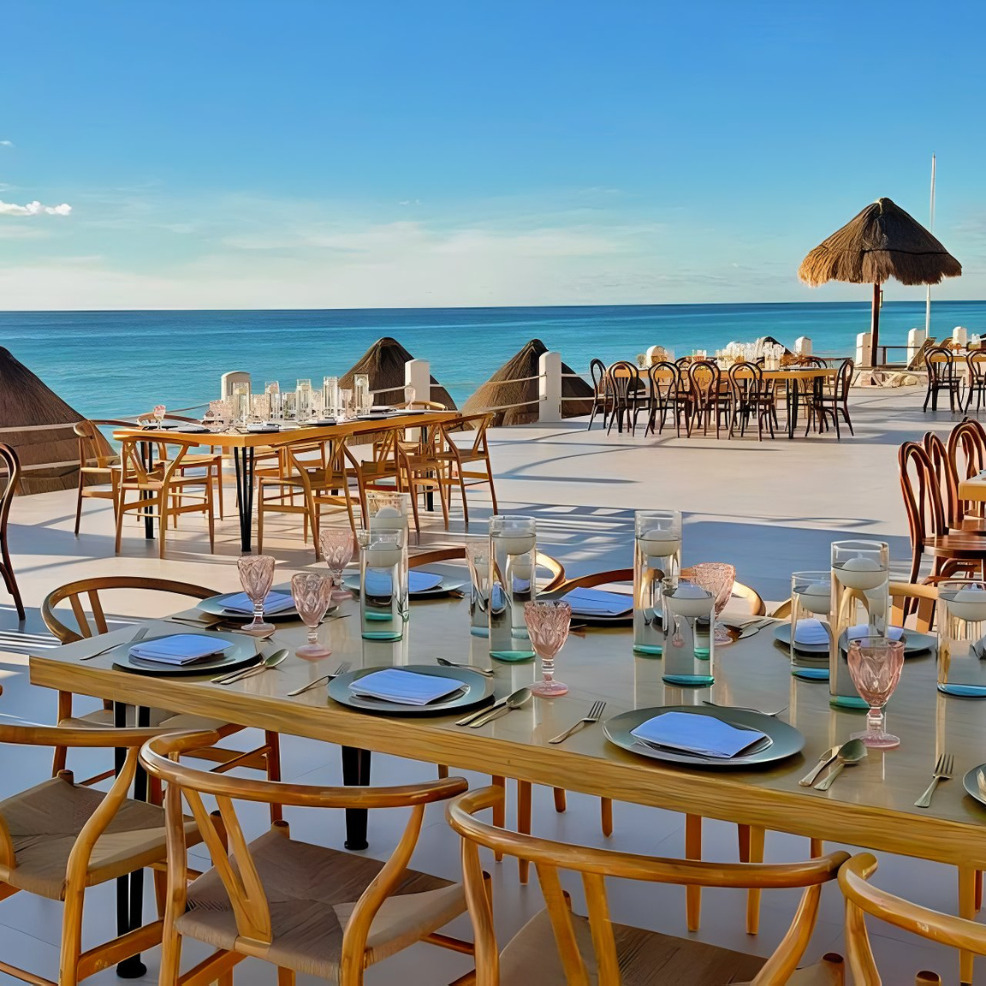 Outdoor dining setup for a destination wedding with several tables set for a meal, overlooking a beach with clear blue sky and straw umbrellas.