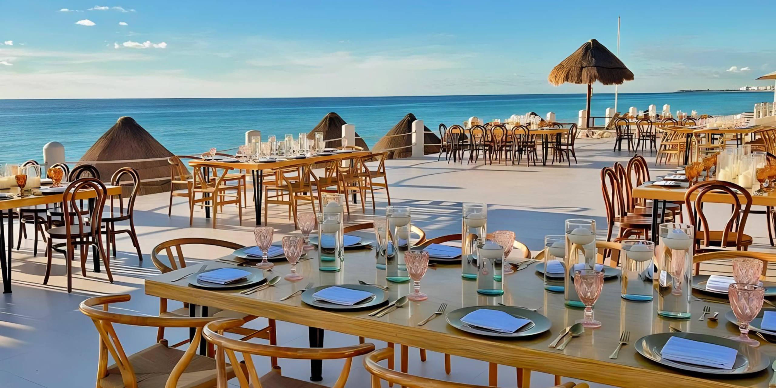 Outdoor beachfront dining area with set tables, wooden chairs, and thatched umbrellas overlooking a calm sea under a clear sky, perfect for a destination wedding.