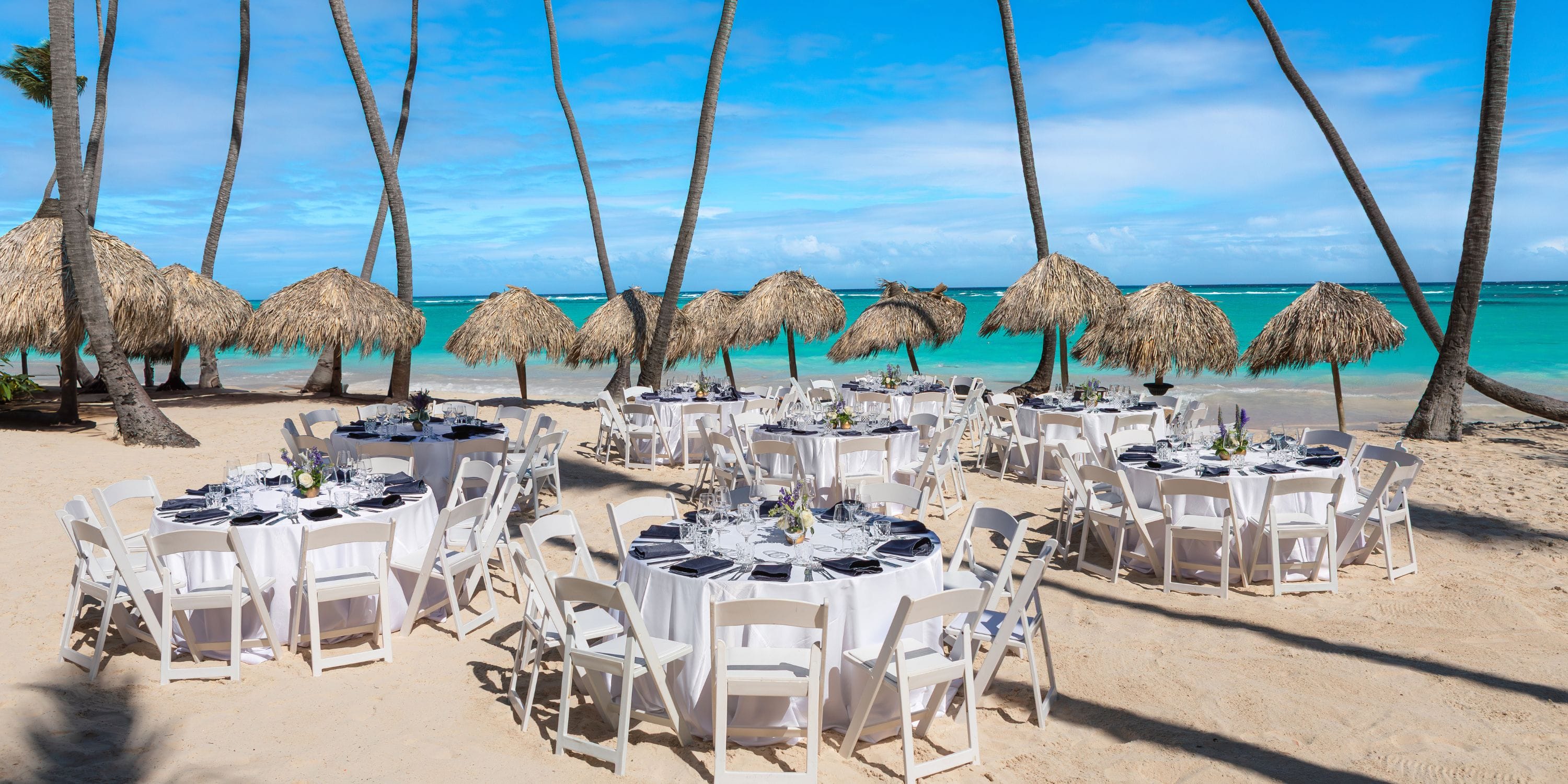 Grand Palladium Select Bavaro: round dining tables set for a destination wedding on a sandy beach with ocean view and palms.