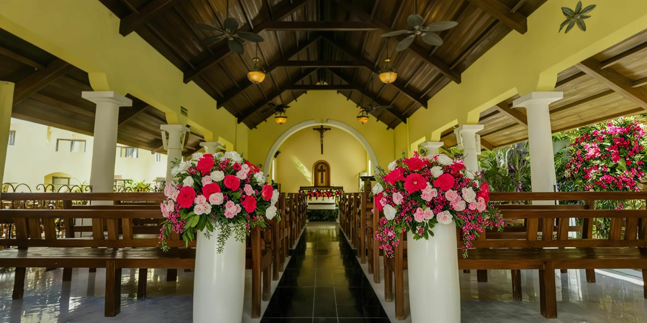 The Grand Palladium Vallarta features a beautifully decorated church interior complete with wooden pews, pink and red flower arrangements lining the aisle, and a crucifix at the altar.