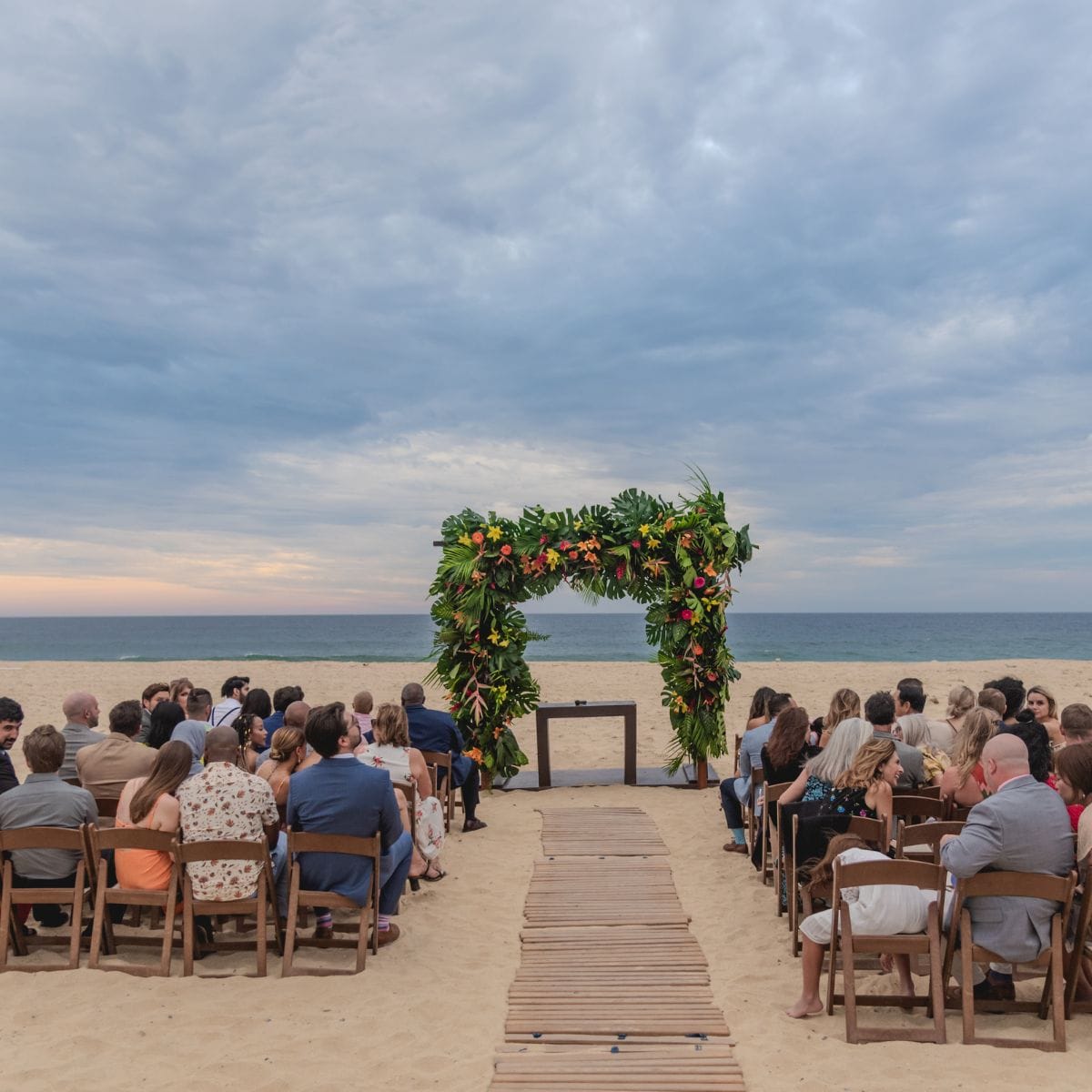 Guests at a destination wedding in Hard Rock Los Cabos face a floral arch and the ocean under clouds.