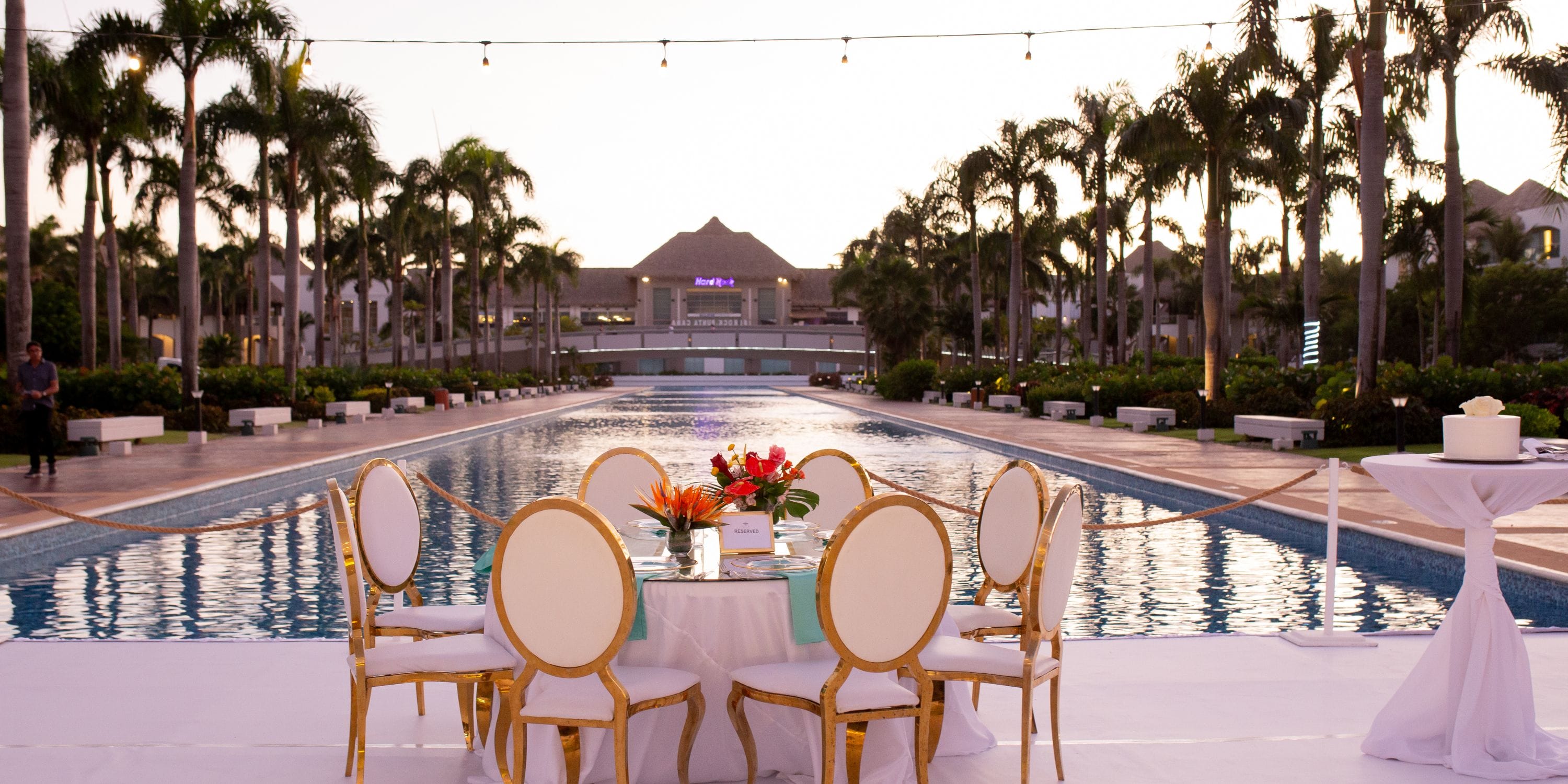 Round wedding table with white chairs by the long pool at Hard Rock Punta Cana, perfect for a destination wedding at sunset among palm trees.