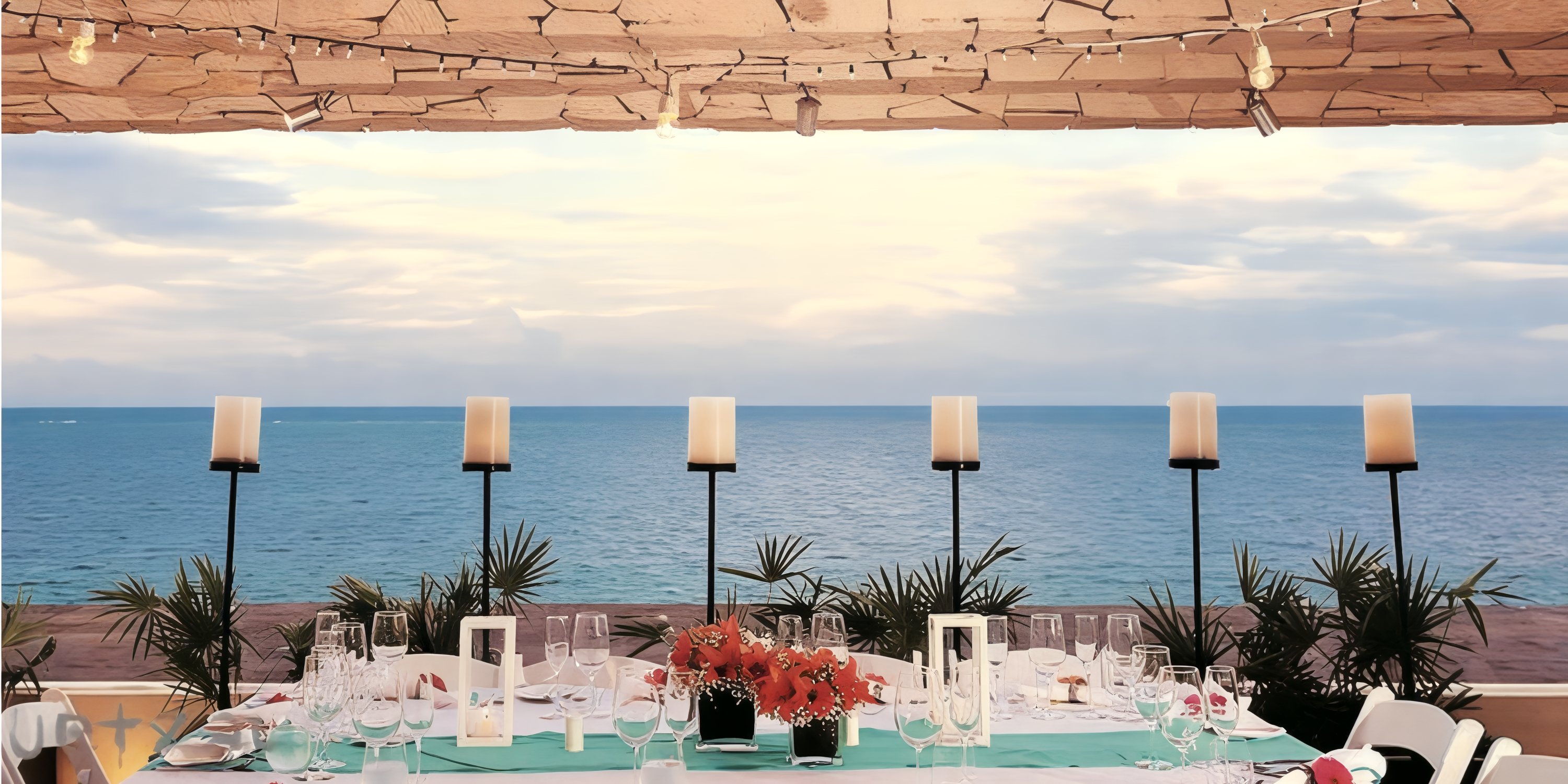 A long dining table set for a destination wedding overlooks the ocean beneath a canopy with string lights.