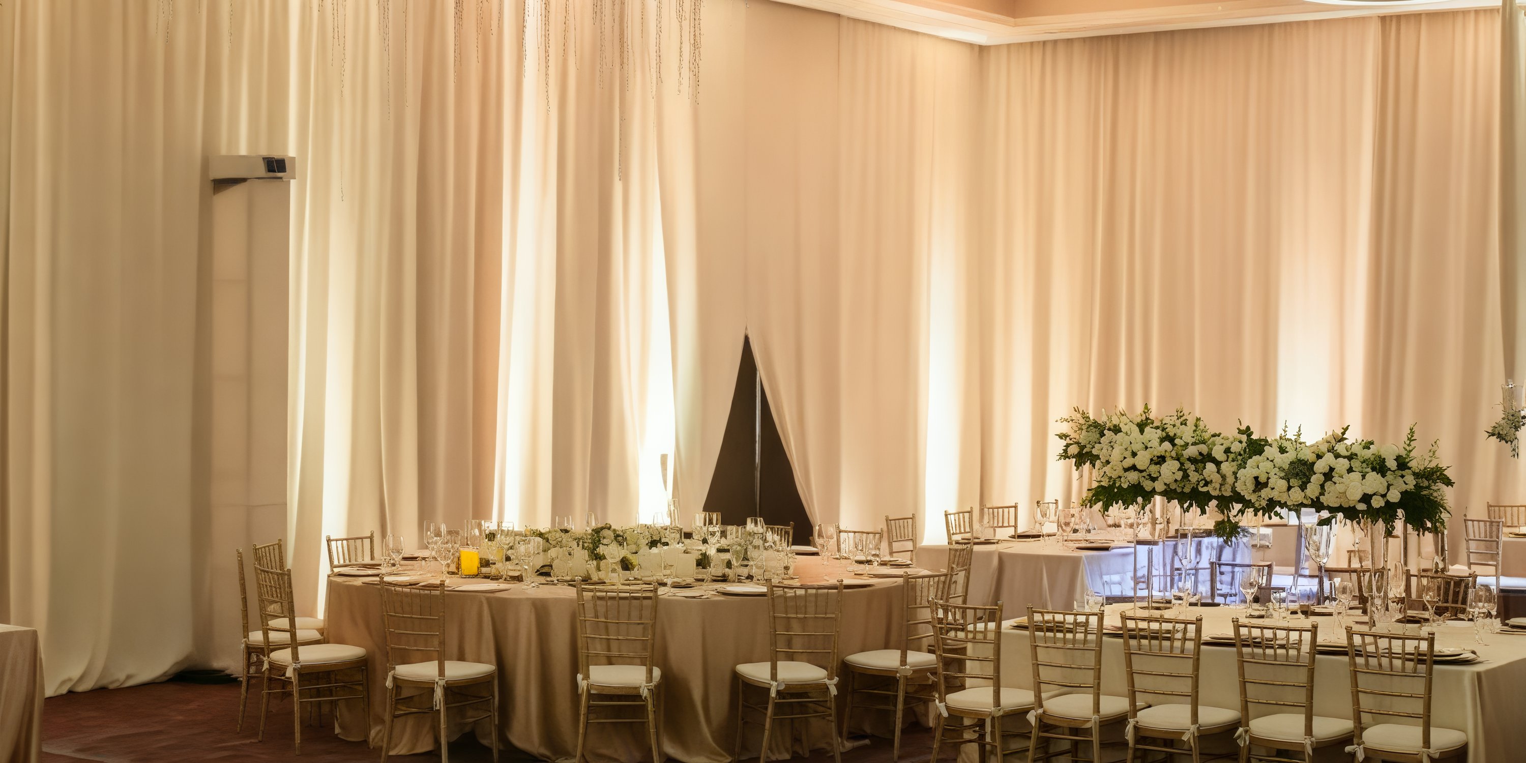 An elegant banquet hall designed for a destination wedding, featuring round tables adorned with white flowers and complemented by tasteful beige drapery.