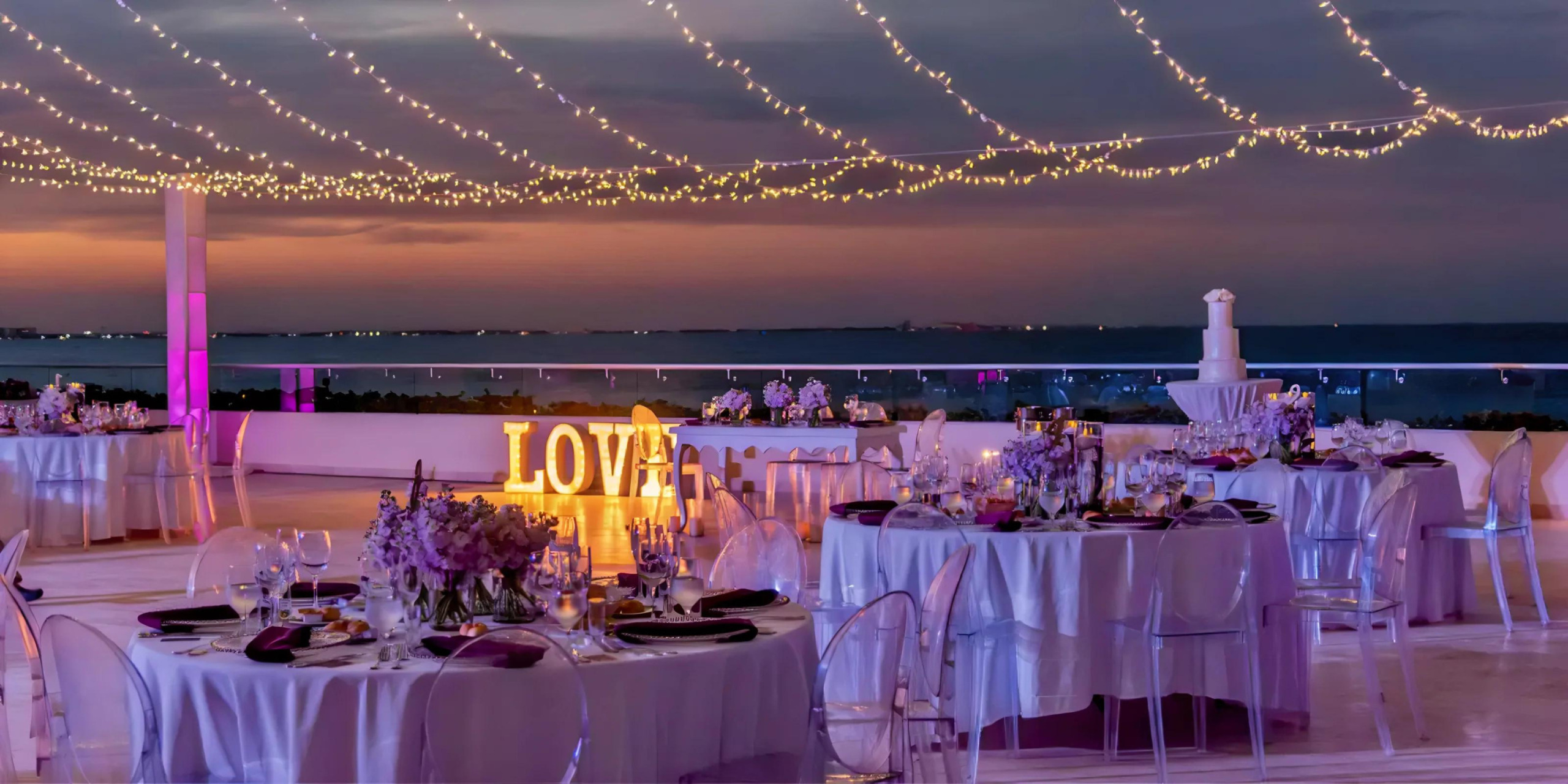 Elegant destination wedding reception at dusk featuring tables set for dinner with purple centerpieces, string lights above, and a "love" sign overlooking a serene water view.