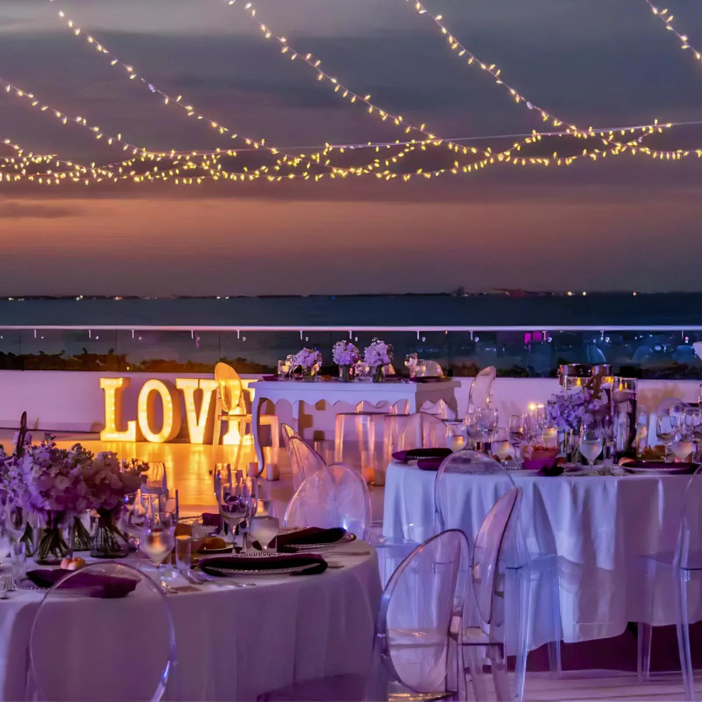 Destination wedding reception at twilight with "love" sign, illuminated string lights, and tables set with flowers under a purple sky.
