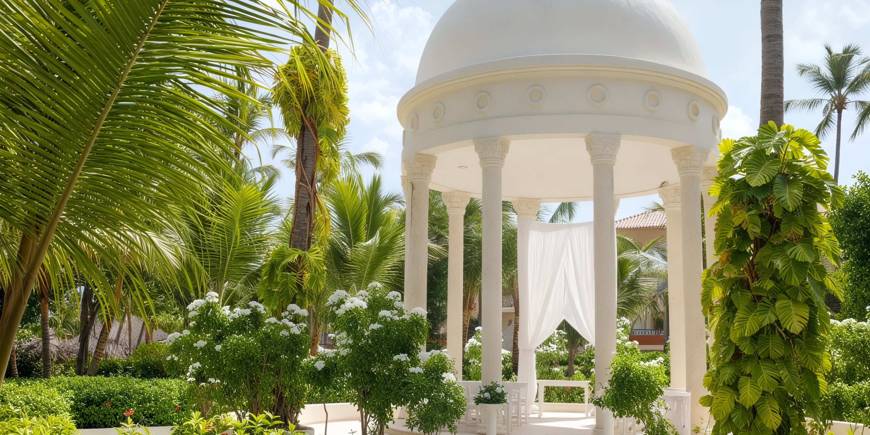 A pristine white gazebo with elegant columns stands amidst lush tropical greenery and swaying palm trees, creating the perfect backdrop for a destination wedding.
