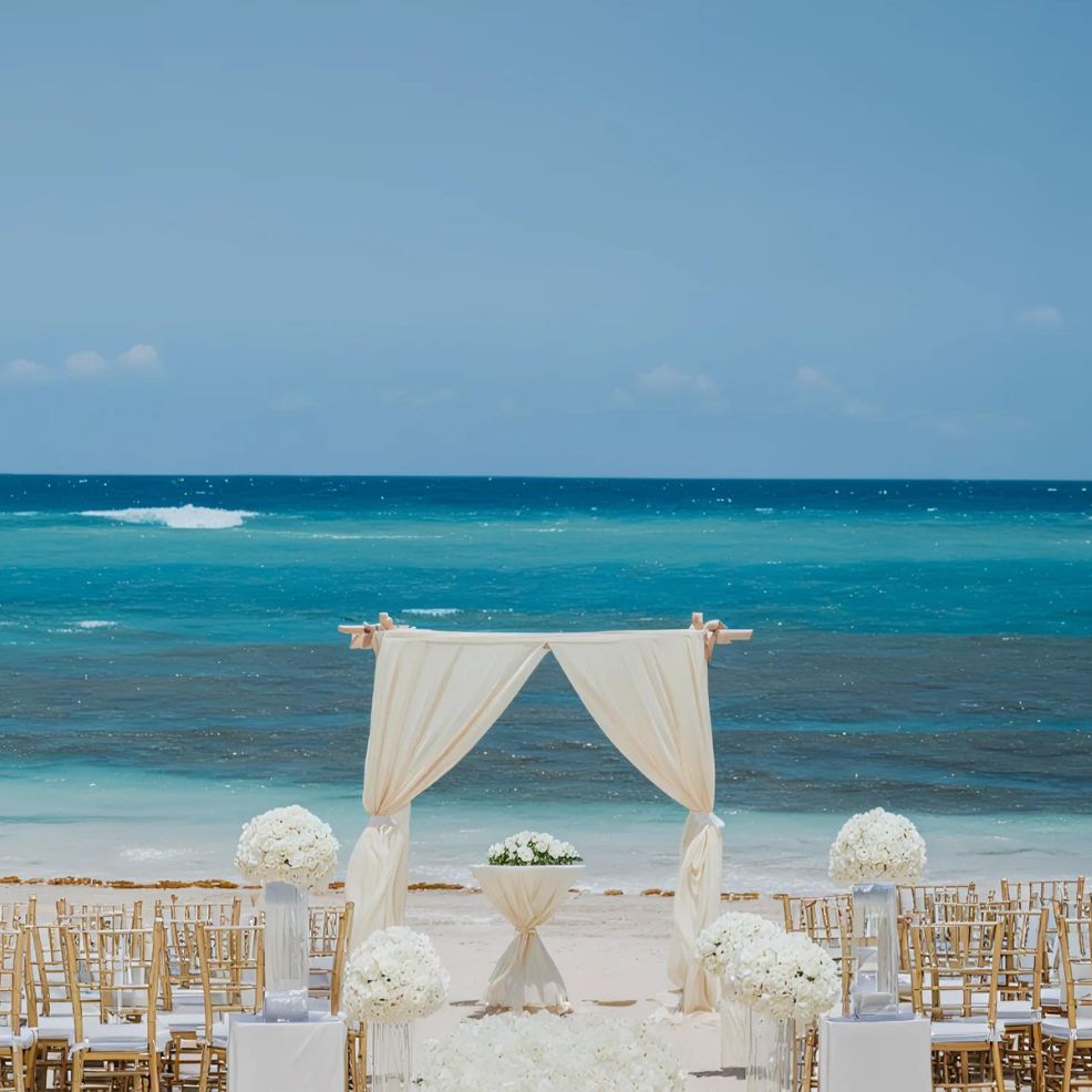 A wedding ceremony setup on a beach, featuring white chairs and floral arrangements, with a white draped arch in front of the ocean and a clear blue sky in the background.