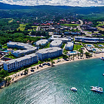 Aerial view of Hotel Riu Montego Bay, popular for destination weddings, with white buildings adjacent to a sandy beach and clear blue sea, with green hills in the background.