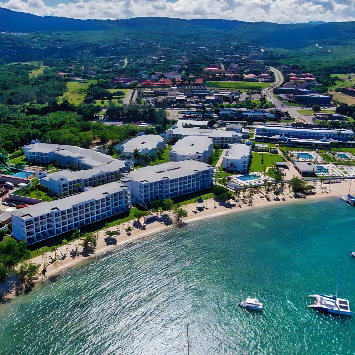 Aerial view of Hotel Riu Montego Bay, popular for destination weddings, with white buildings adjacent to a sandy beach and clear blue sea, with green hills in the background.
