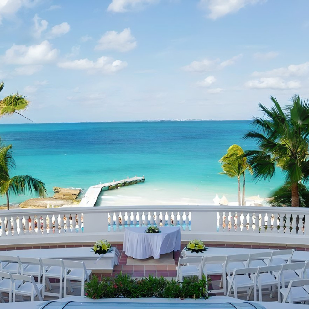 Tropical beach view from a balcony set up for destination weddings, featuring white chairs, table with a white cloth, and a clear blue ocean backdrop.