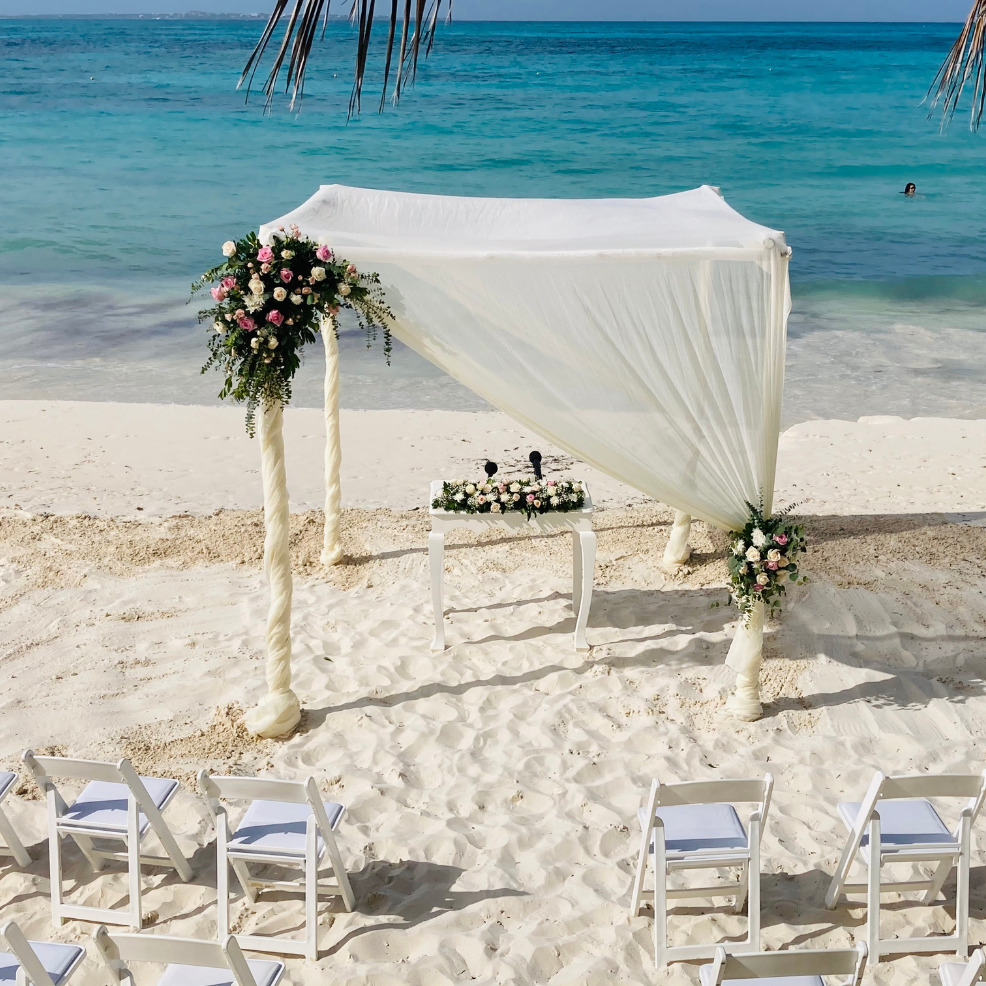 A destination beach wedding setup with a white canopy decorated with flowers, white chairs arranged on the sand, and a clear blue ocean in the background.